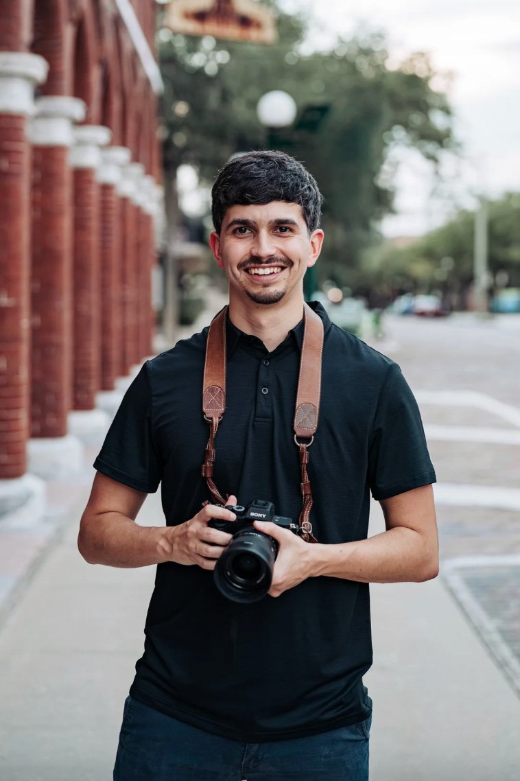 A young man with dark hair, a goatee, and a smile, standing outdoors on a sidewalk. He is holding a camera in his hands and wearing a black polo shirt with a leather strap around his neck, likely attached to the camera. The background includes a brick building with white columns and blurred trees.