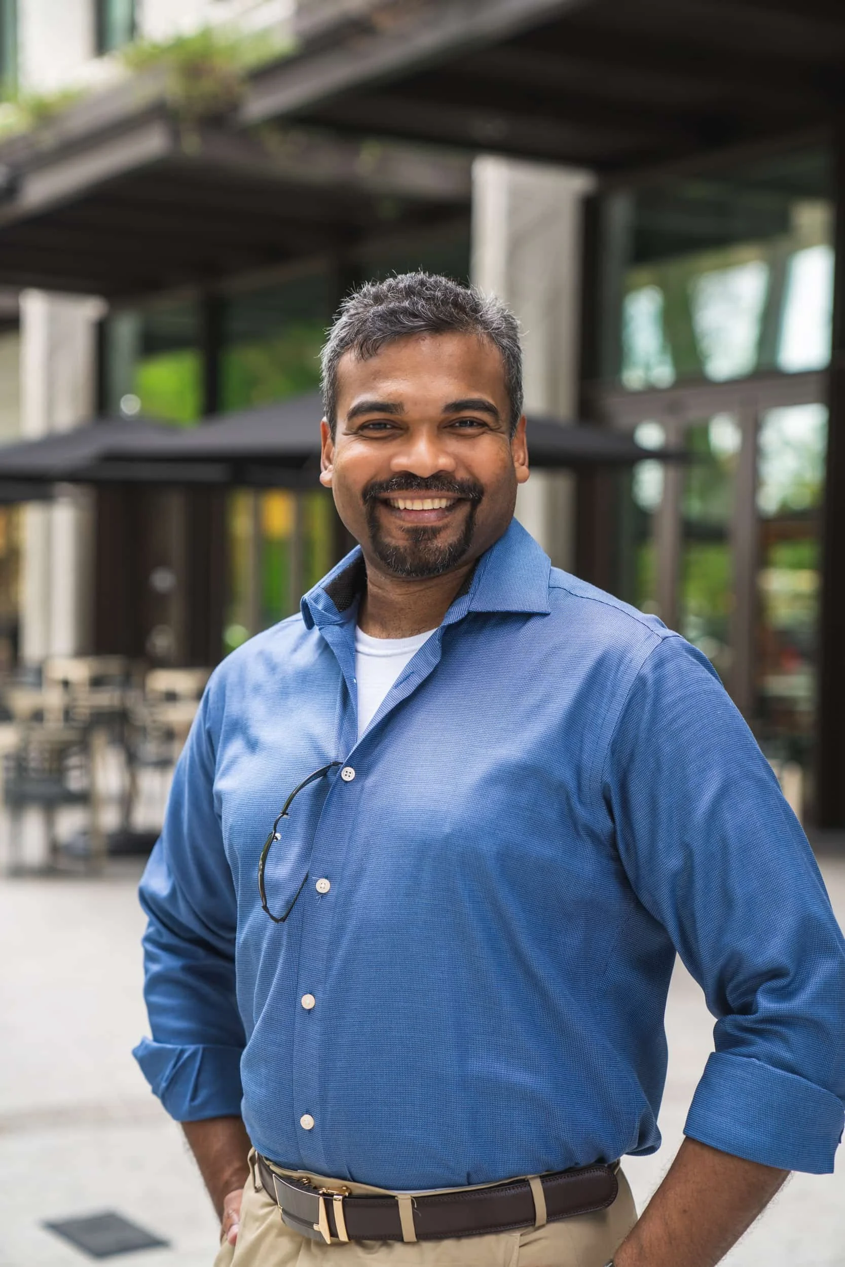 Smiling man with short gray hair, goatee, wearing a blue button-up shirt and beige pants, outdoors with modern architecture and greenery in the background.