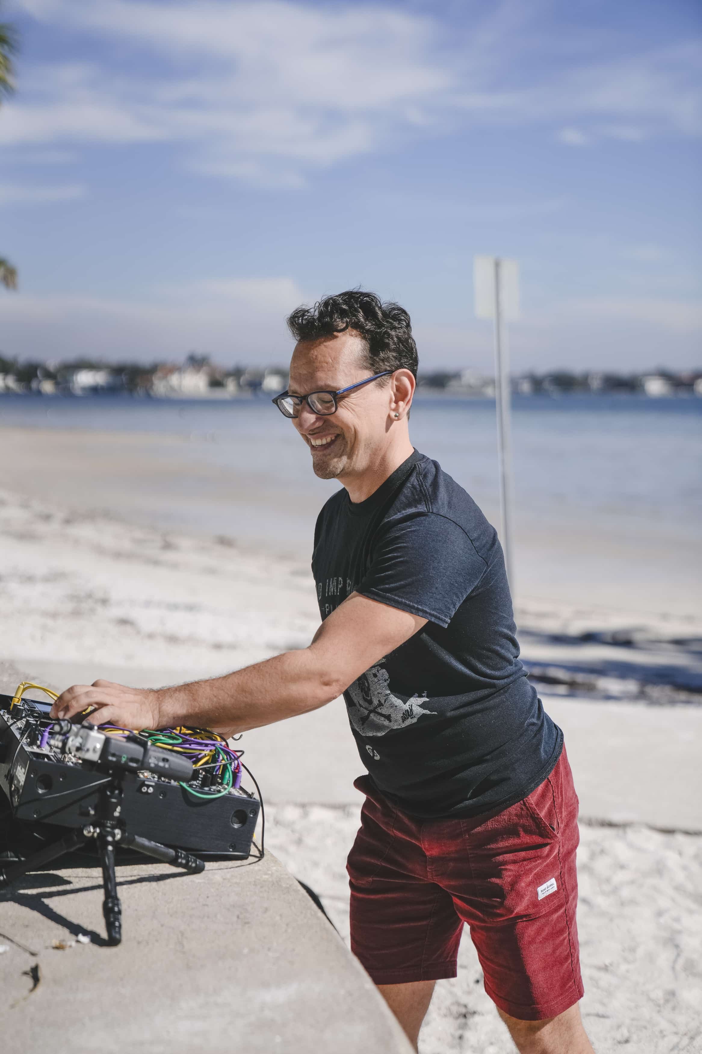 A man with glasses and a big smile interacting with electronic music equipment on a beach, with water and houses in the background on a sunny day.