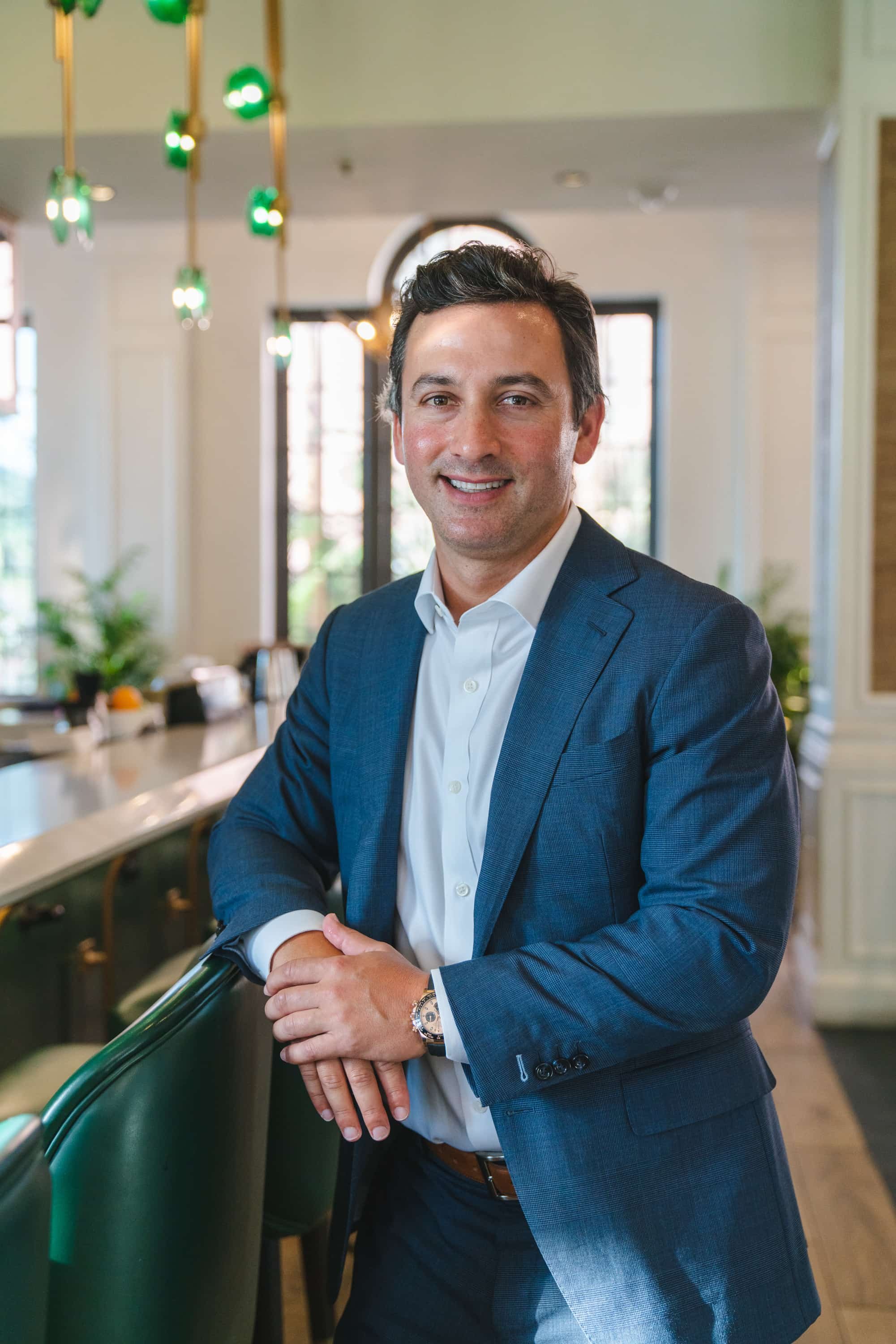 A man in a navy blue suit and white shirt stands in a restaurant, leaning on a green banquette, smiling at the camera. The background shows large windows and hanging green lights.