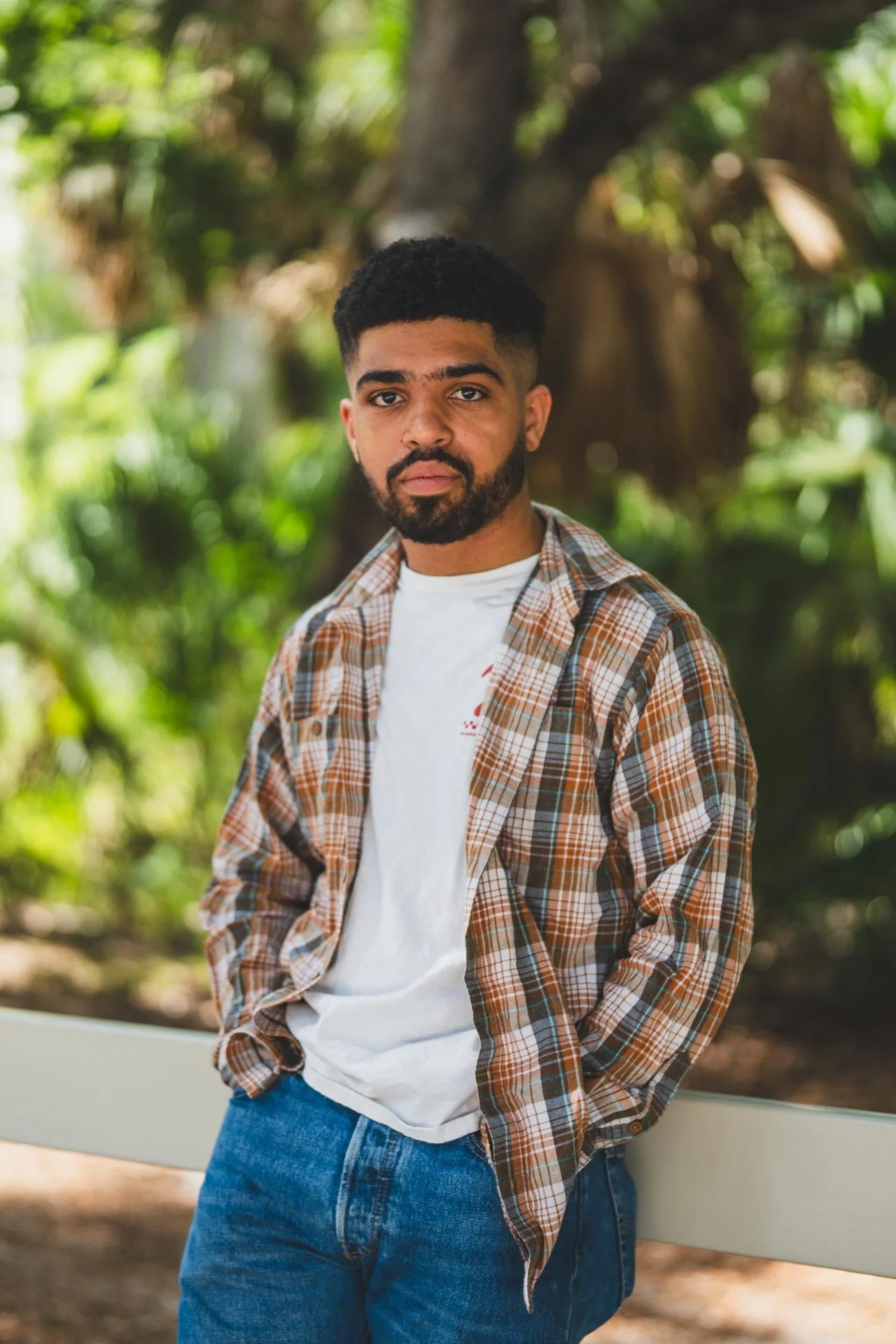 A young man with a beard and short curly hair, wearing a plaid shirt over a white t-shirt and blue jeans, standing outdoors in front of a tree and greenery, looking at the camera with a neutral expression.