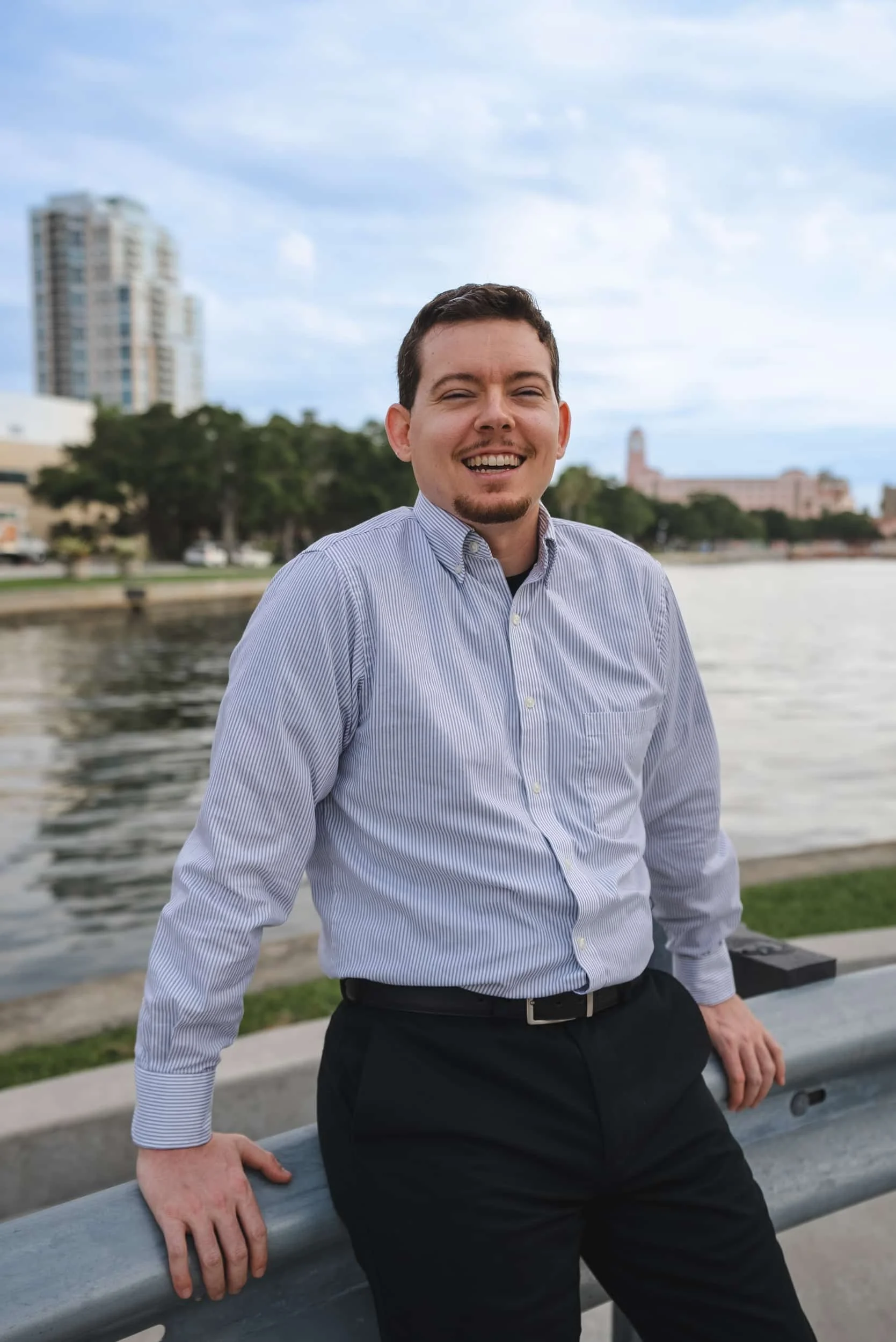 A young man with short dark hair, a goatee, wearing a striped button-up shirt and black pants, standing by a river with city buildings and trees in the background. He is smiling and leaning on a metal railing.
