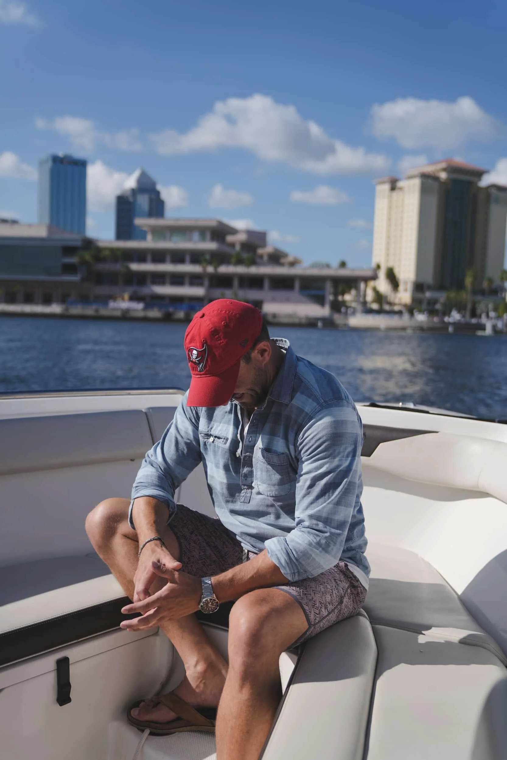 A man sitting on a boat, wearing a red Cap with a skull logo, sunglasses, a blue checkered shirt, and shorts, with a city skyline and water in the background.