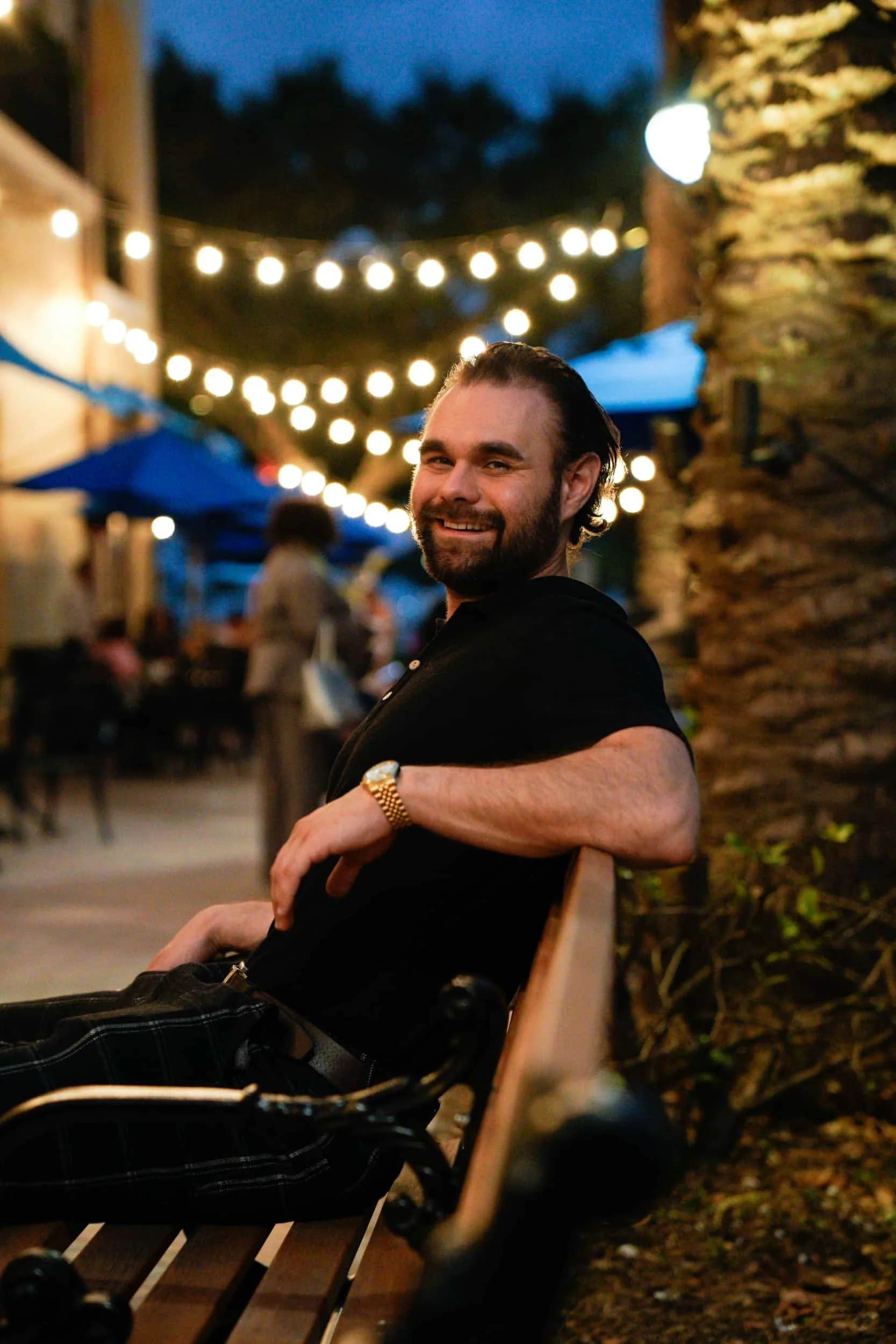 A smiling man with dark hair and beard sitting on a bench outdoors at night, with string lights hanging above him and a large tree trunk to his right, in a lively area with people and blue umbrellas in the background.