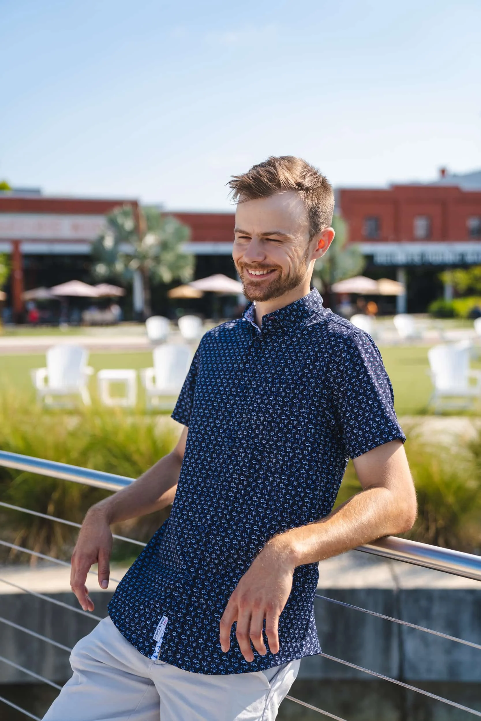 A young man with short hair and a beard smiling outdoors on a sunny day, wearing a short-sleeved patterned shirt and light-colored pants, leaning on a metal railing with a blurred background of outdoor seating and a red brick building.