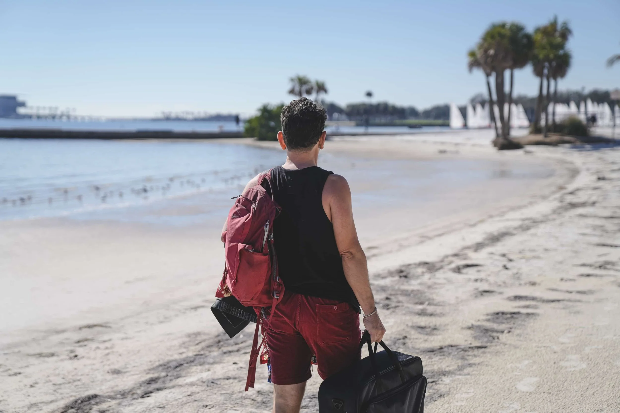 A person with short dark hair wearing a black tank top and red shorts, carrying a red backpack and a black duffel bag, walking on a sandy beach near the ocean with trees and sailboats in the background.