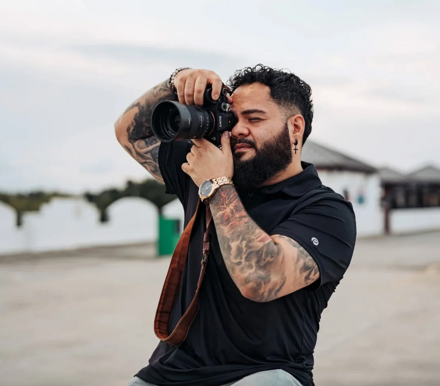 A man with tattoos and a beard is taking a photograph with a Sony camera outdoors during daytime. He is wearing a black shirt, a gold watch, and a beaded bracelet, and appears to be focusing intently with his eye close to the camera viewfinder.