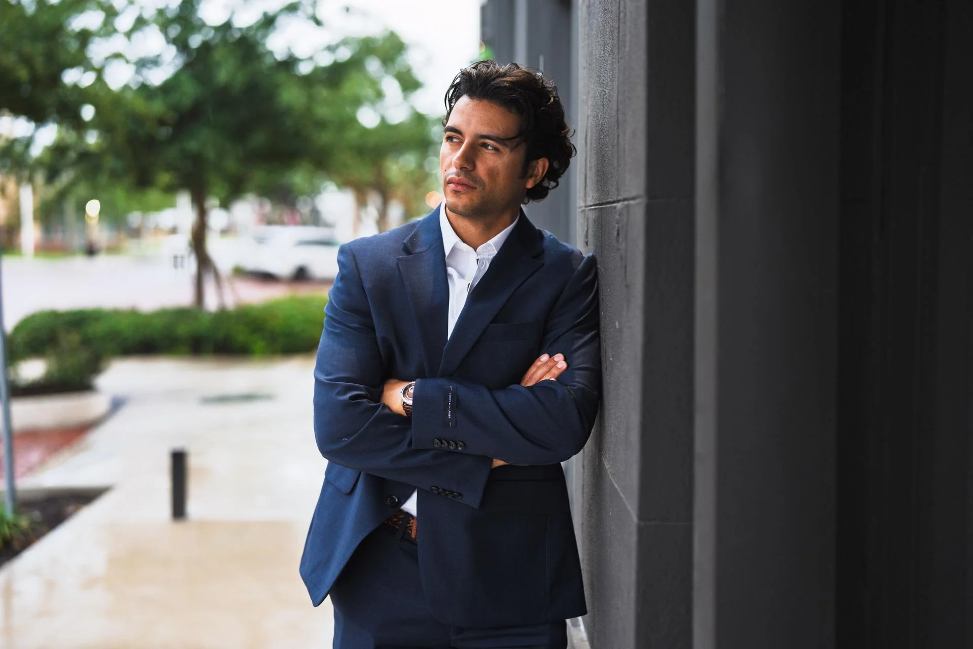 Man in a navy blue suit with crossed arms leaning against a dark wall outdoors.