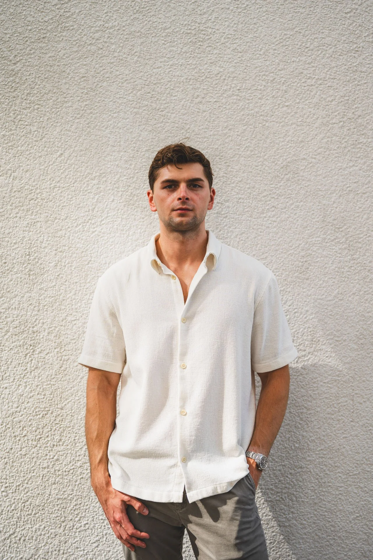 A young man with short brown hair, wearing a cream-colored short-sleeve button-up shirt and dark pants, standing against a textured white wall.