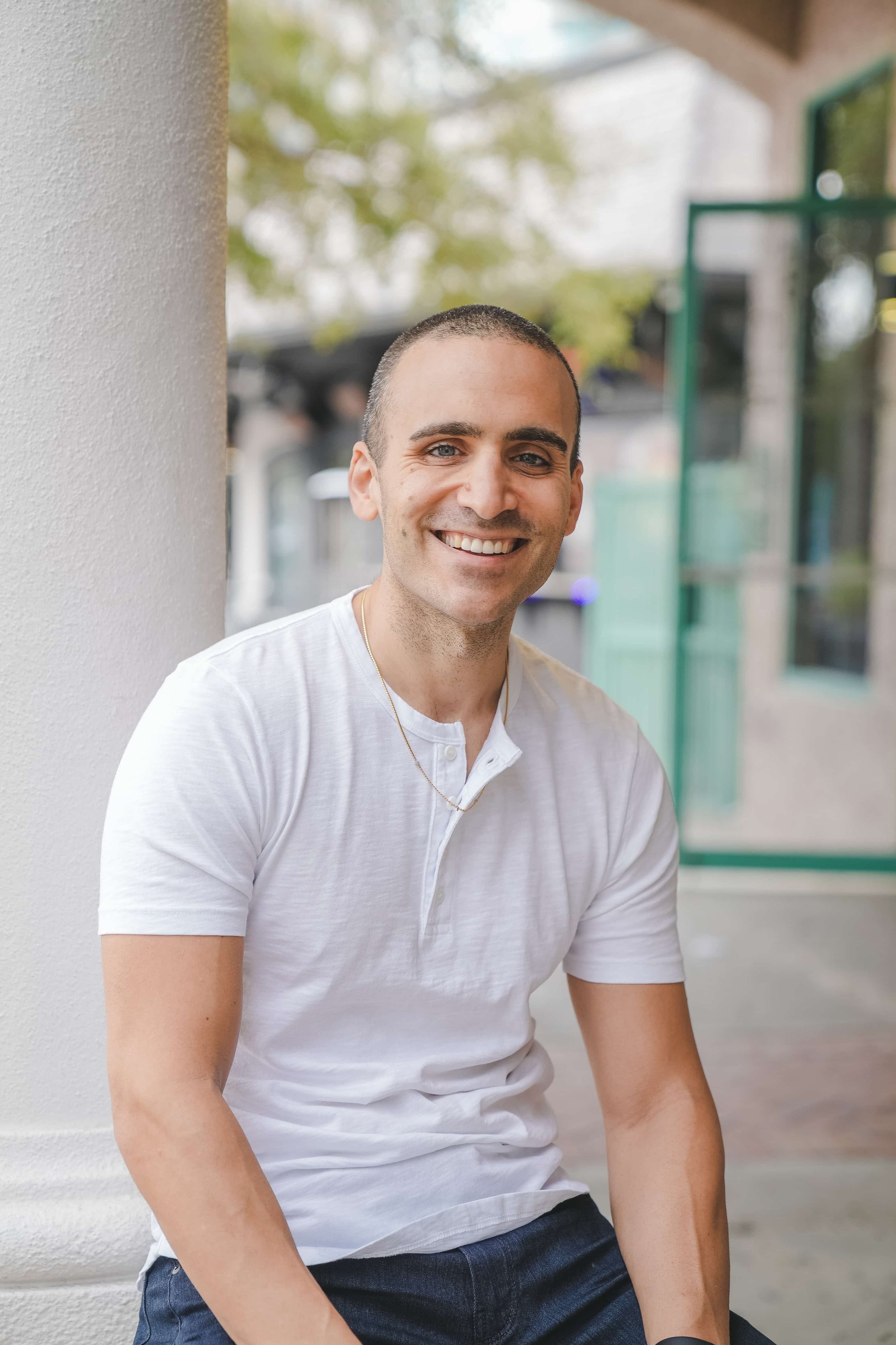A smiling man with short dark hair, wearing a white t-shirt and dark jeans, sitting outdoors near a white wall with a blurred background of trees and glass windows.