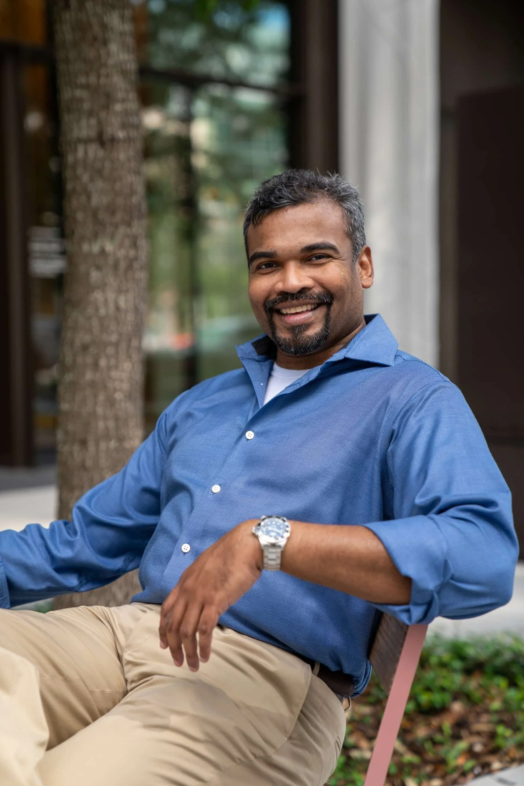 A smiling man with dark hair and a beard, wearing a blue button-up shirt and beige pants, is sitting on a pink outdoor chair with a watch on his left wrist. He is outdoors, with trees and a building in the background.