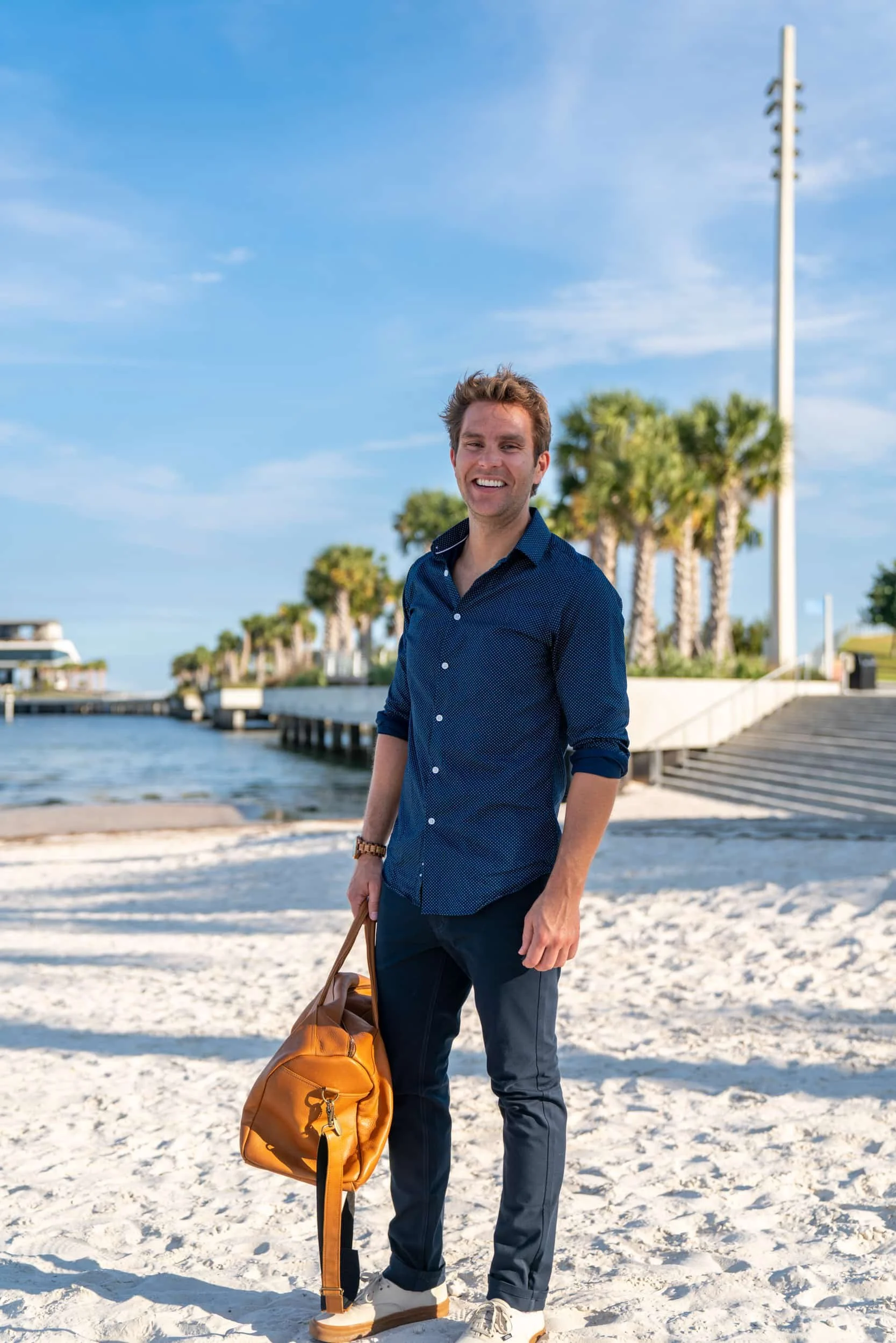 A man standing on a sandy beach holding a brown duffle bag, smiling at the camera with palm trees, a pier, and boats in the background on a sunny day.