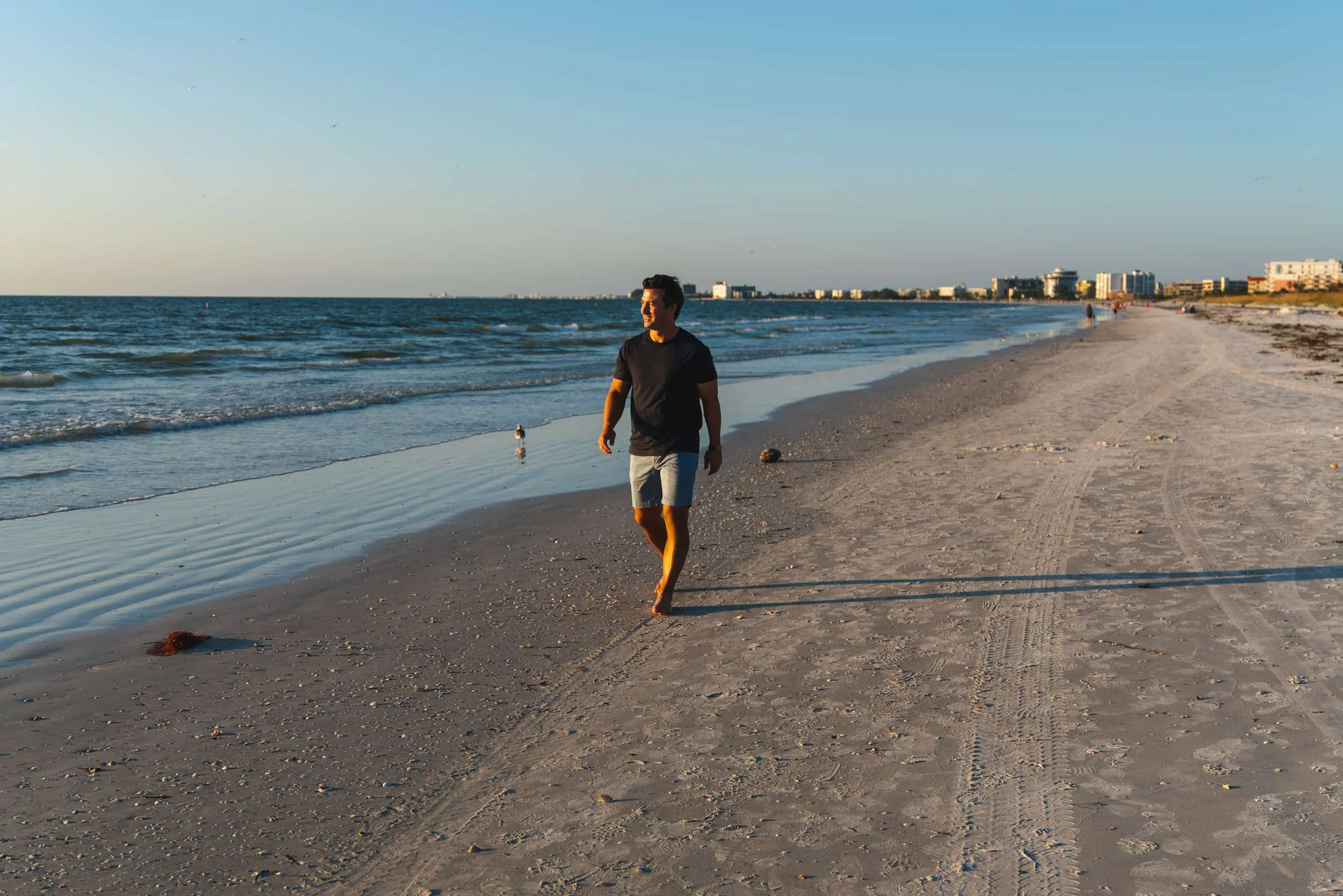 A man walking barefoot along the sandy shoreline of a beach with buildings in the background, the sky clear and blue.