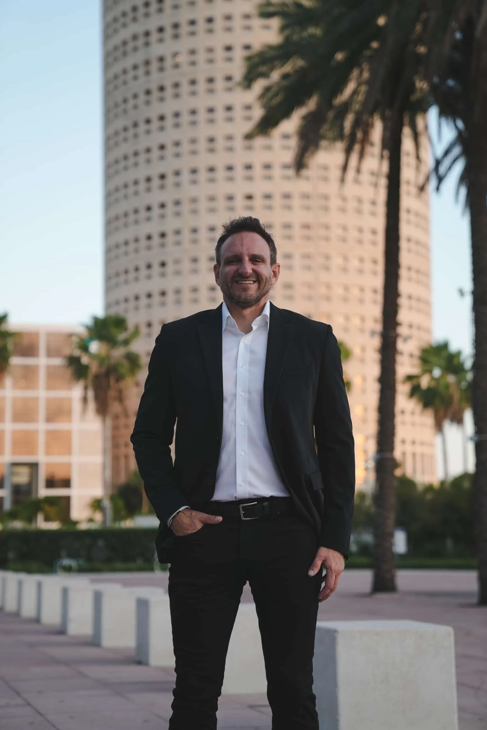 A man in a black suit and white shirt standing outdoors with a modern high-rise building and palm trees in the background, smiling in the evening