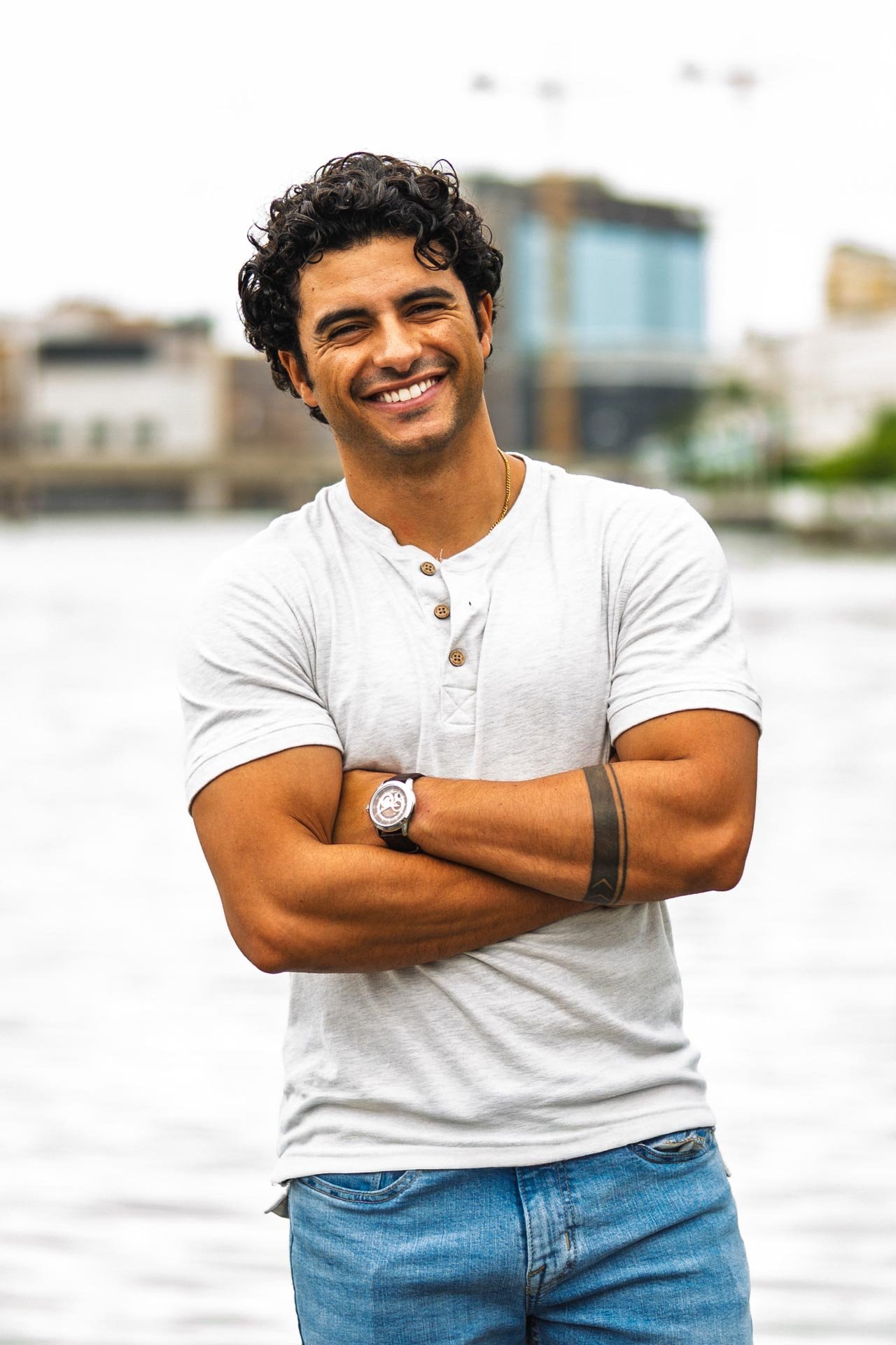 A man with curly black hair and a dark tan skin tone standing outdoors during daytime, smiling with his arms crossed, wearing a white t-shirt, blue jeans, a wristwatch, and a necklace with a small pendant, with a blurred background of buildings and w
