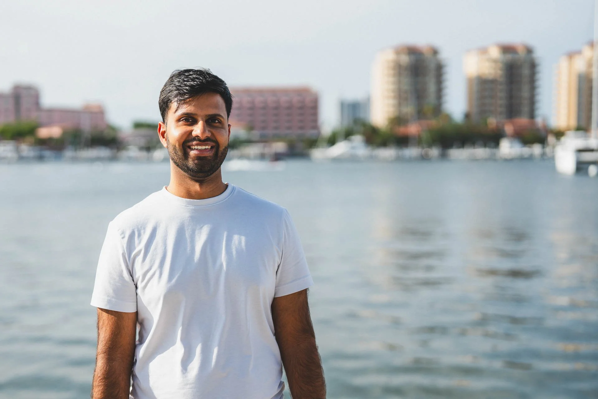 A smiling man with a beard and short dark hair in a white t-shirt standing by a body of water with city buildings in the background.