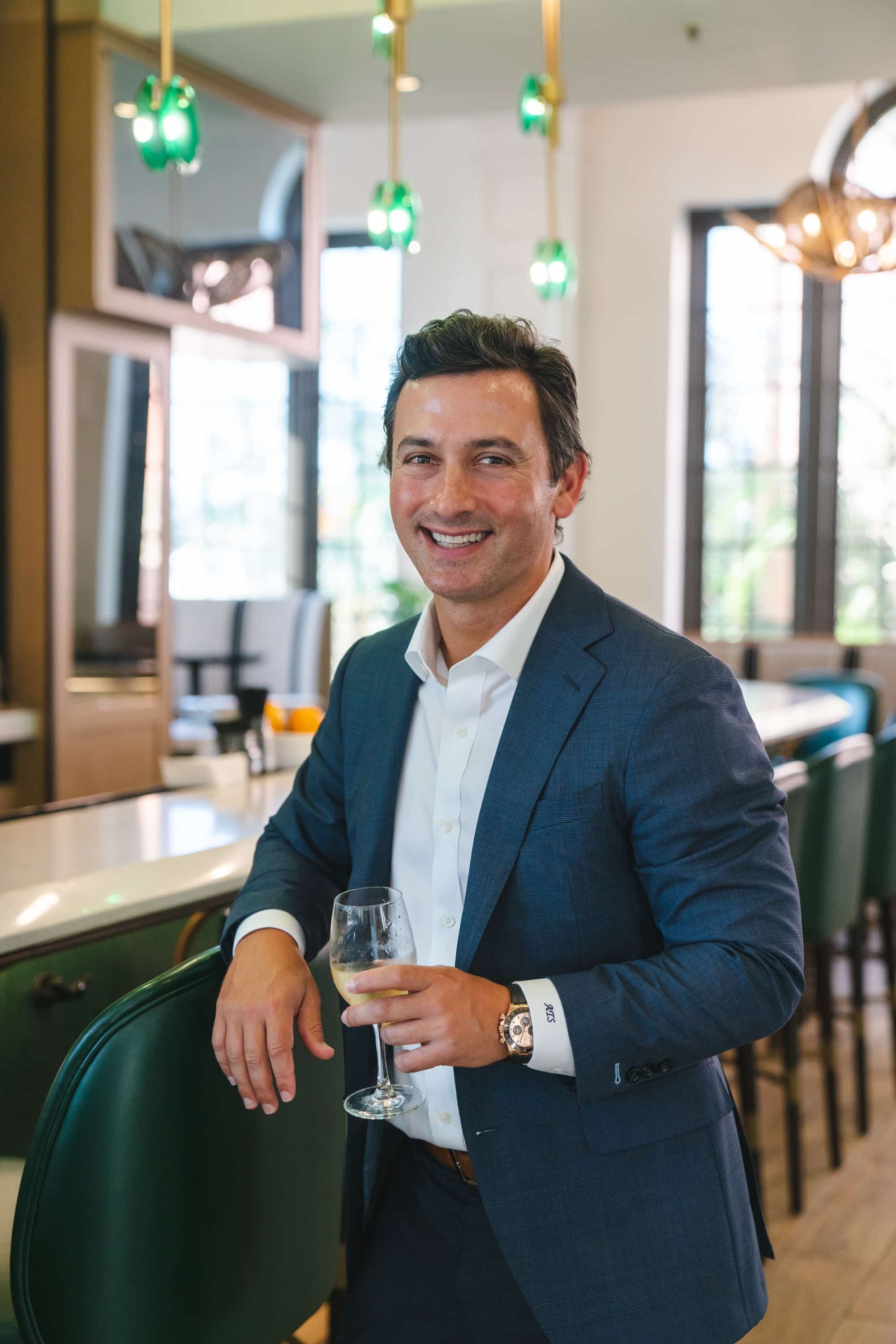 A man in a blue suit holding a glass of white wine in a restaurant or bar setting, smiling at the camera.