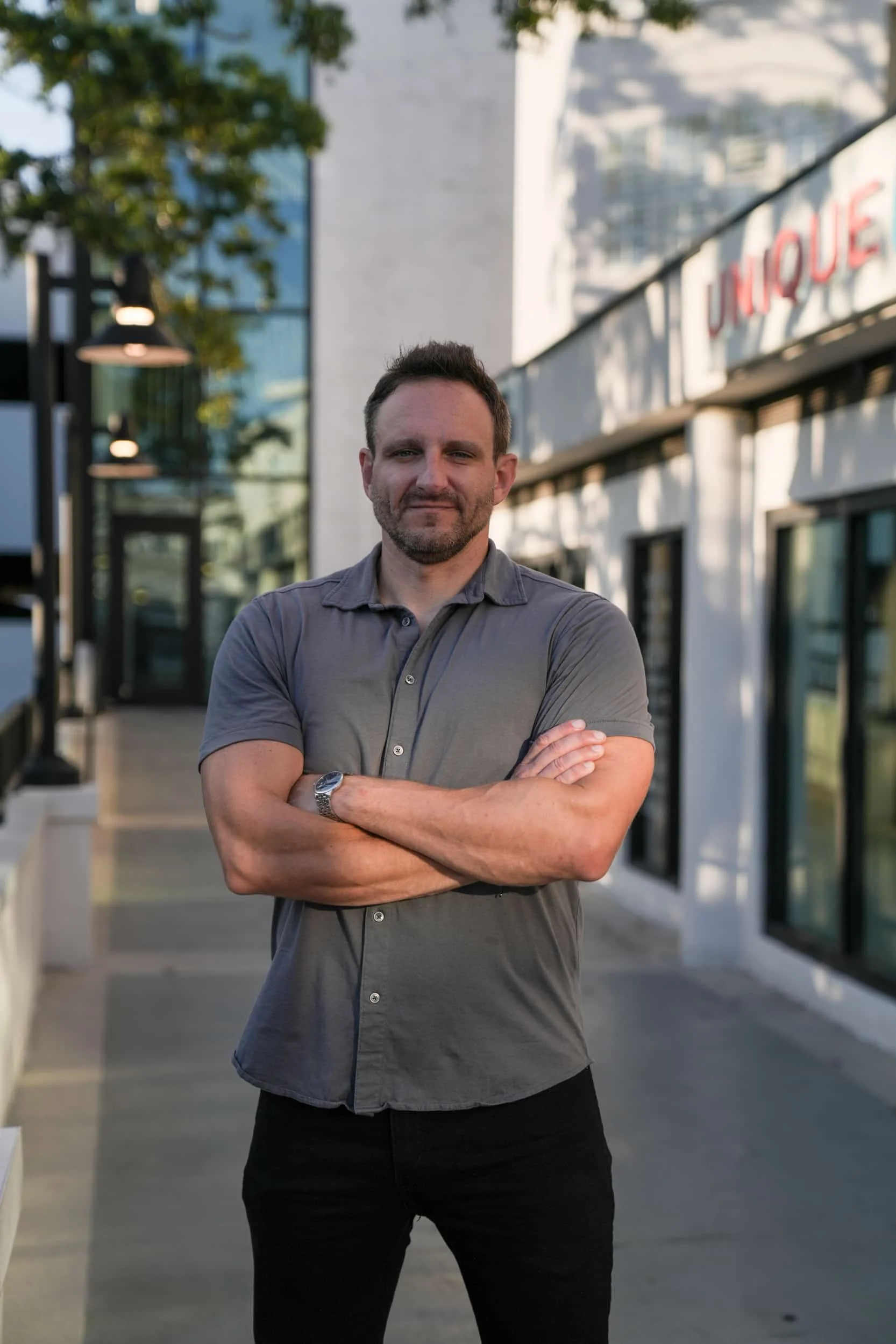 A man with short dark hair and a beard stands outdoors with arms crossed, wearing a gray button-up shirt and black pants, in front of a modern building with large glass windows and a sign that reads 'JUNIQUE' on a white wall.