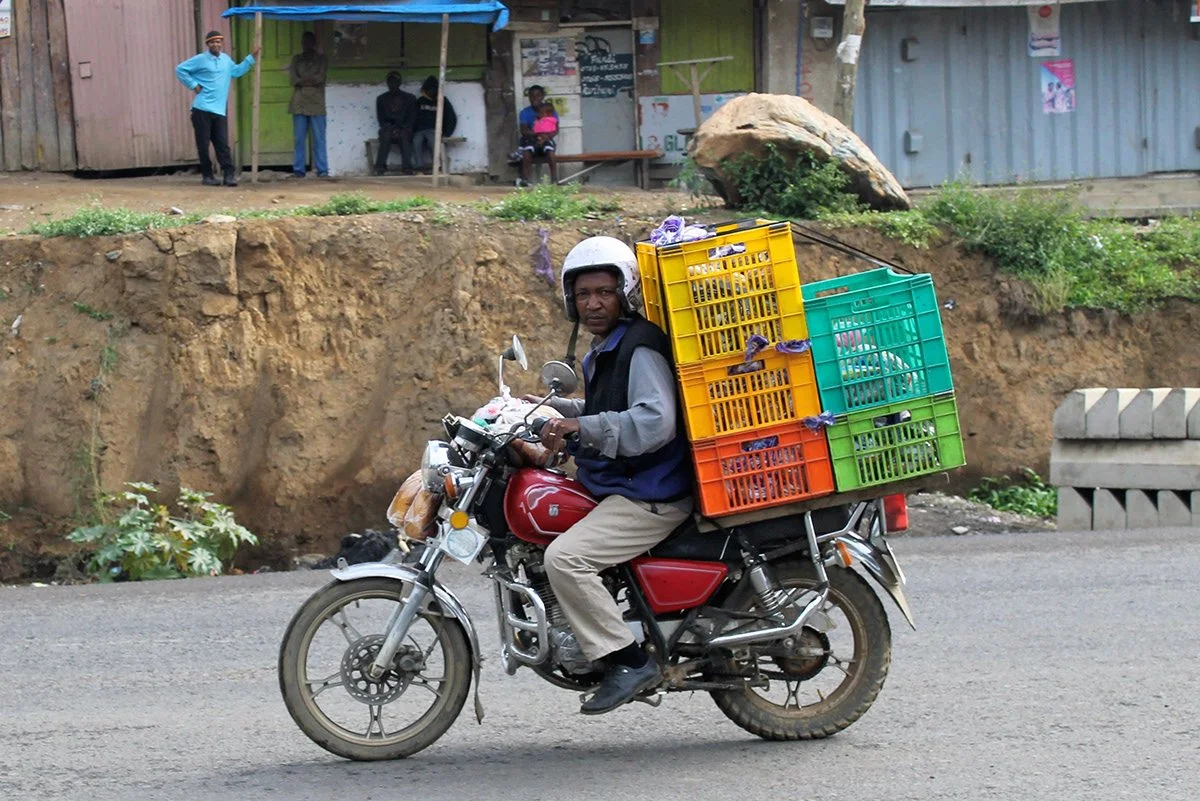 A man wearing a helmet riding a motorcycle on a paved road with plastic crates loaded on the back, in a rural or semi-urban area.