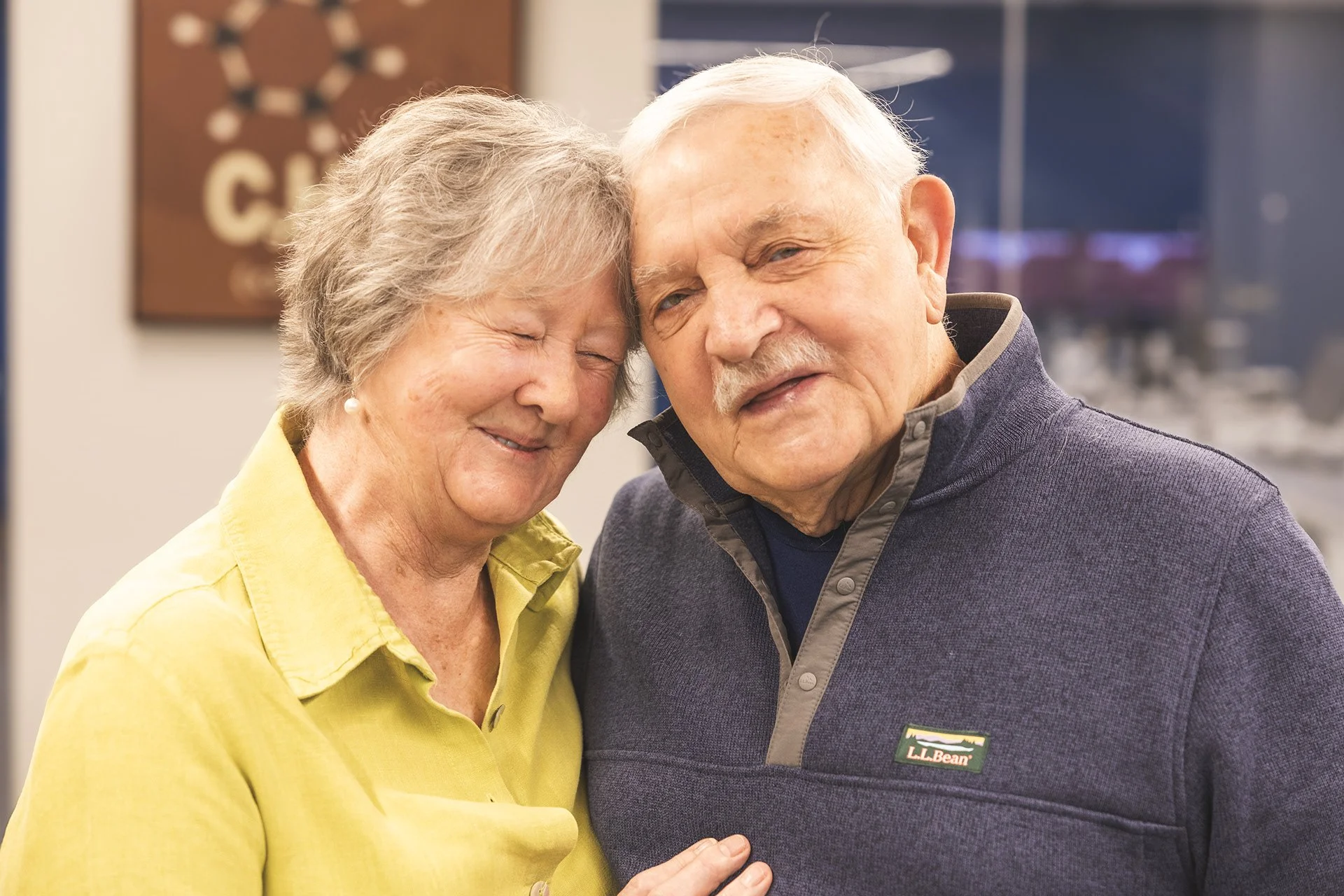 An elderly woman with gray hair smiling with her eyes closed, standing close to an elderly man with white hair and a mustache, both are dressed casually and appear to be happy.