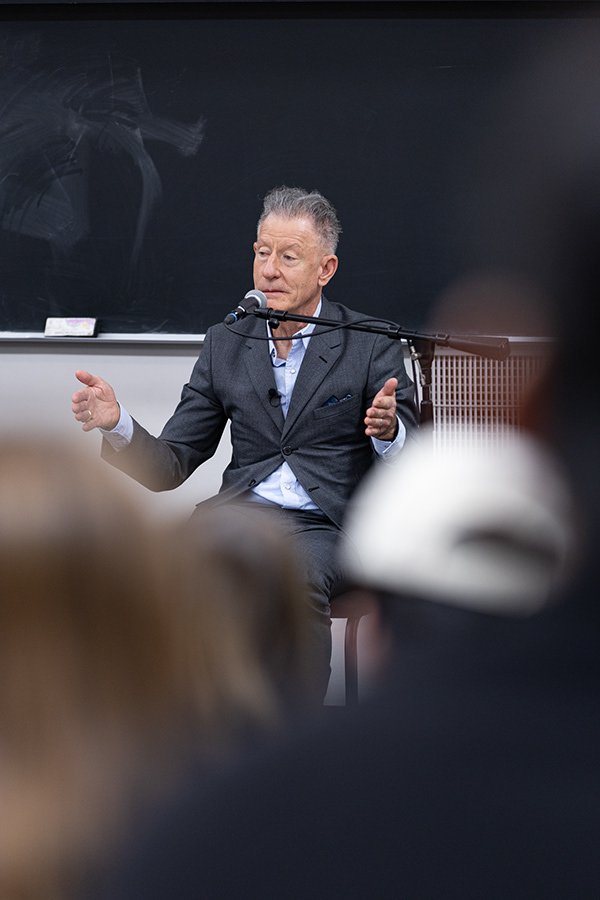 A man in a suit sitting in front of a blackboard speaks into a microphone during a lecture or discussion, with an audience in the foreground.