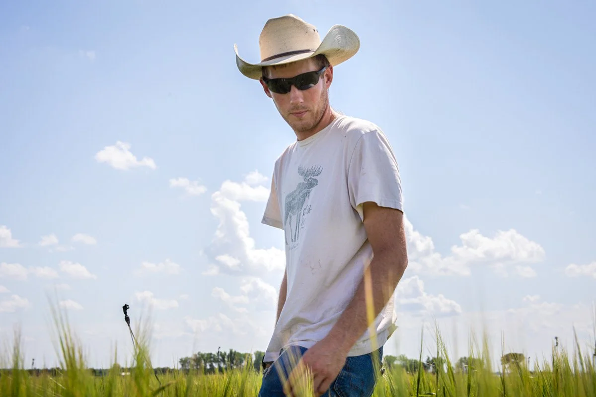 A man wearing a cowboy hat and sunglasses standing in a grassy field on a sunny day.