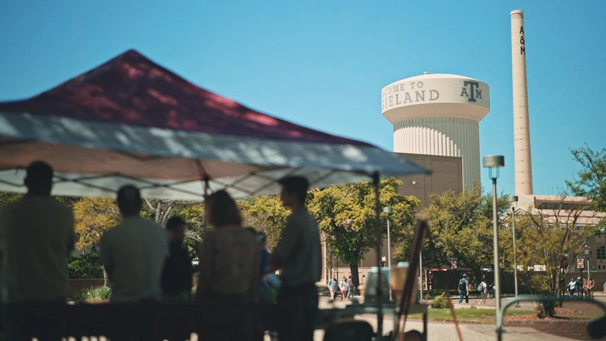 People under a tent in front of the Texas A&M University campus with the iconic A&M Tower in the background.