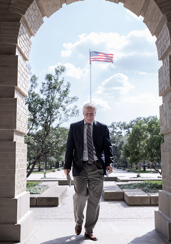 An elderly man in a suit walking out of a stone archway, holding a book and a smartphone, with an American flag in the background and trees in a park.
