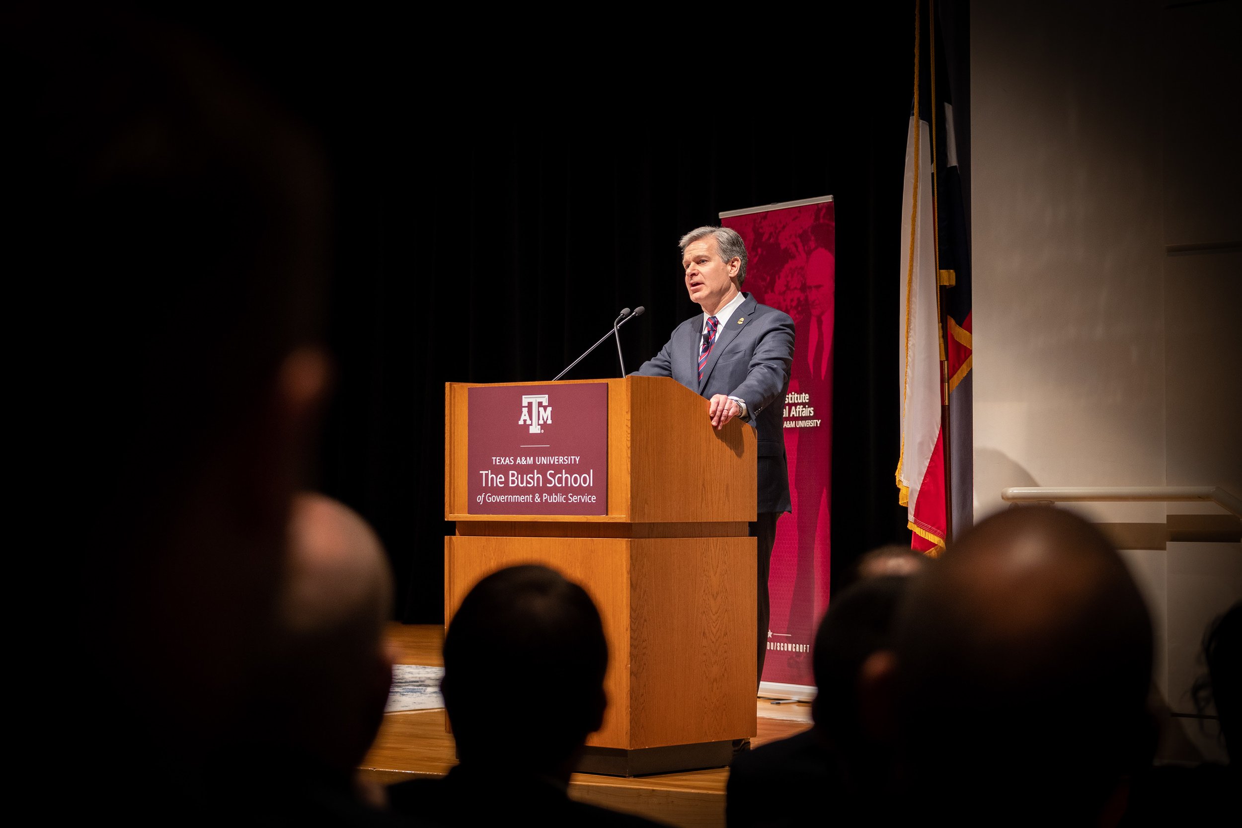 A man in a suit speaking at a podium with a Texas A&M University banner and flags behind him, audience in foreground.