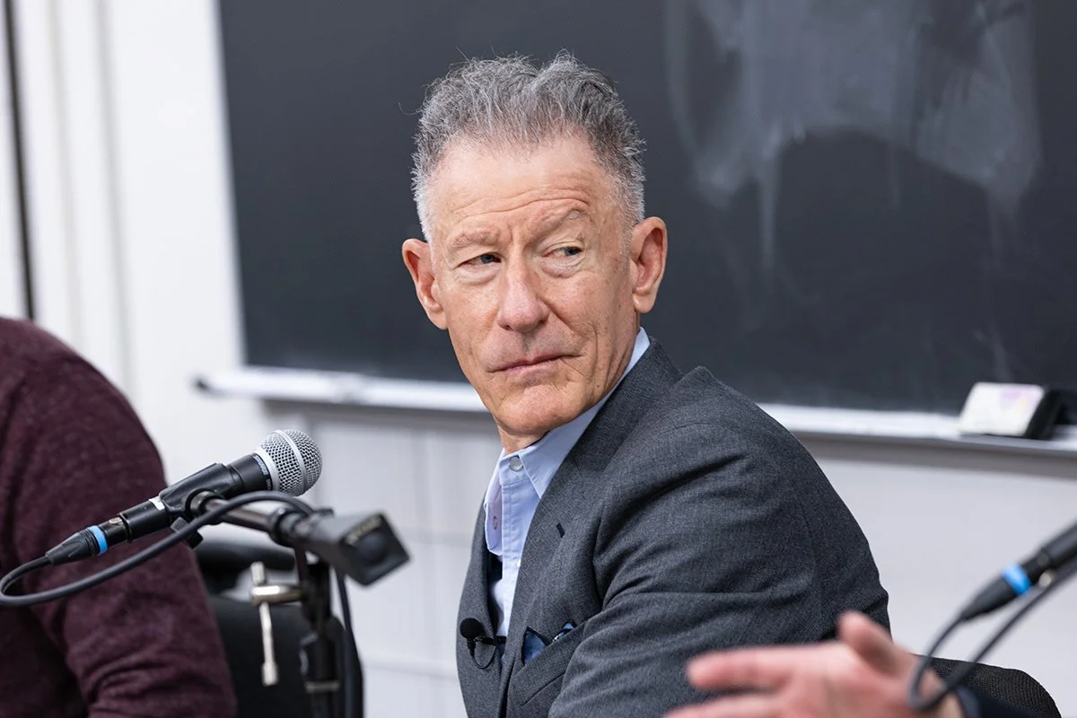 An older man in a gray suit and light blue shirt looking to the right, sitting at a table with a microphone in front of him, in a classroom or conference room setting.