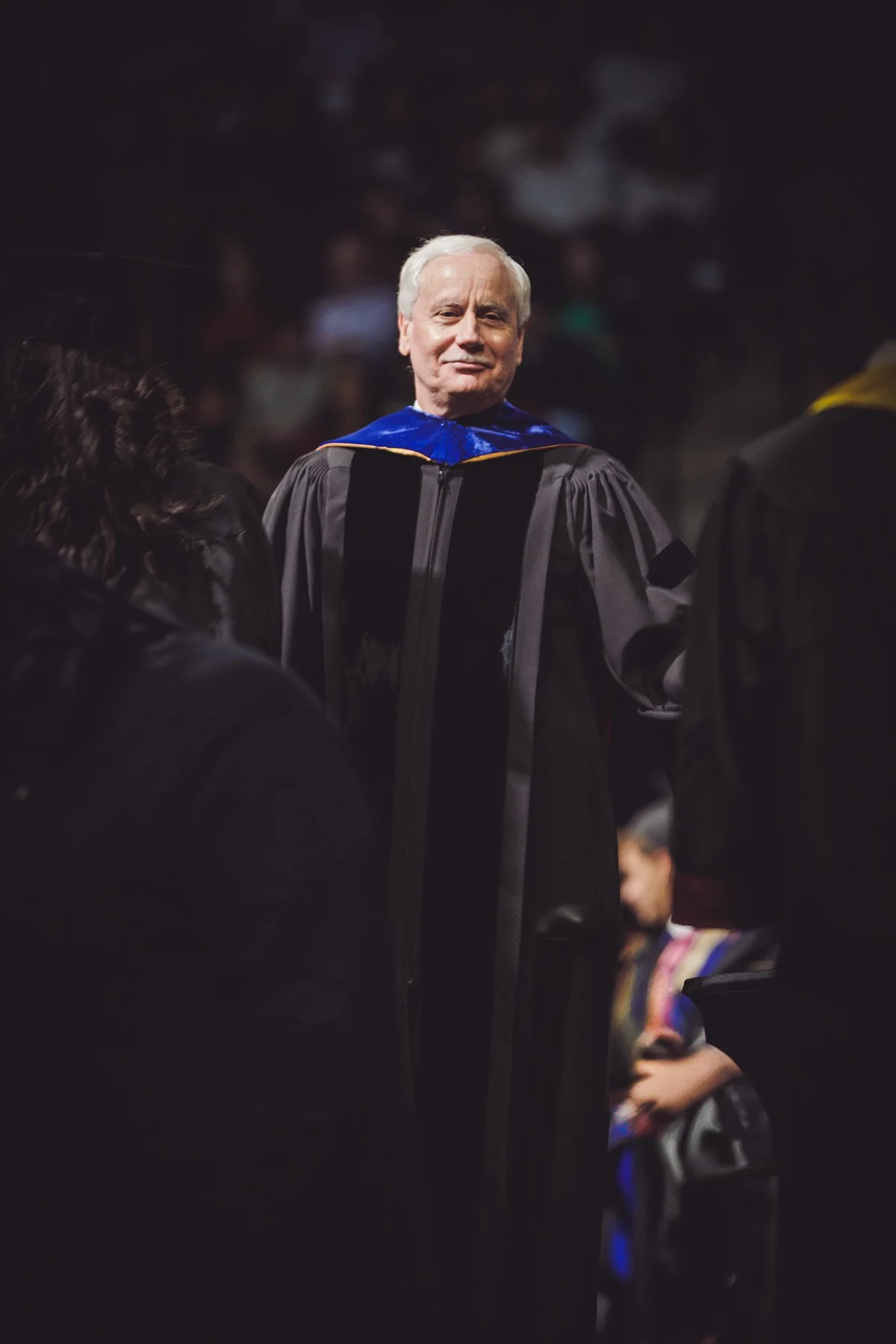 An elderly man in academic regalia, including a black gown and a blue hood, standing at a graduation ceremony. He is smiling and appears to be in a large, dark auditorium with other graduates and attendees.