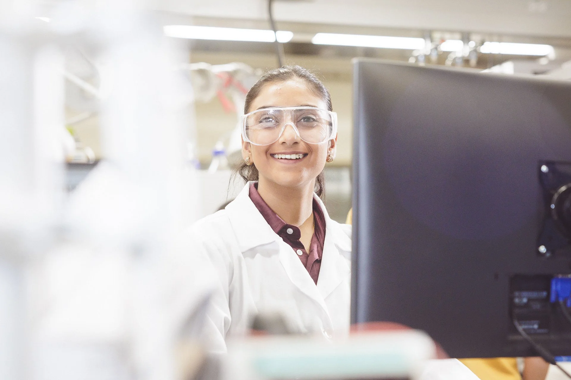 A smiling woman in a laboratory wearing safety goggles and a white lab coat, working at her computer with lab equipment in the background.
