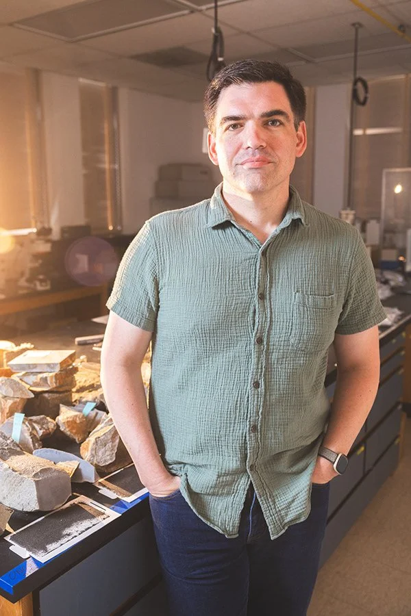 A man with dark hair and dressed in a green short-sleeve shirt and dark jeans, standing in a laboratory or workshop with rocks and equipment on a black counter.