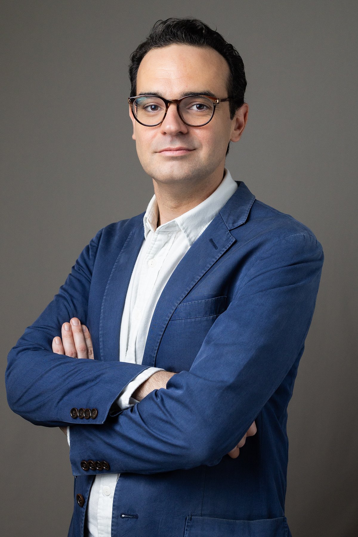 Portrait of a man with dark curly hair, wearing glasses, a white shirt, and a blue suit jacket, standing with arms crossed against a gray background.