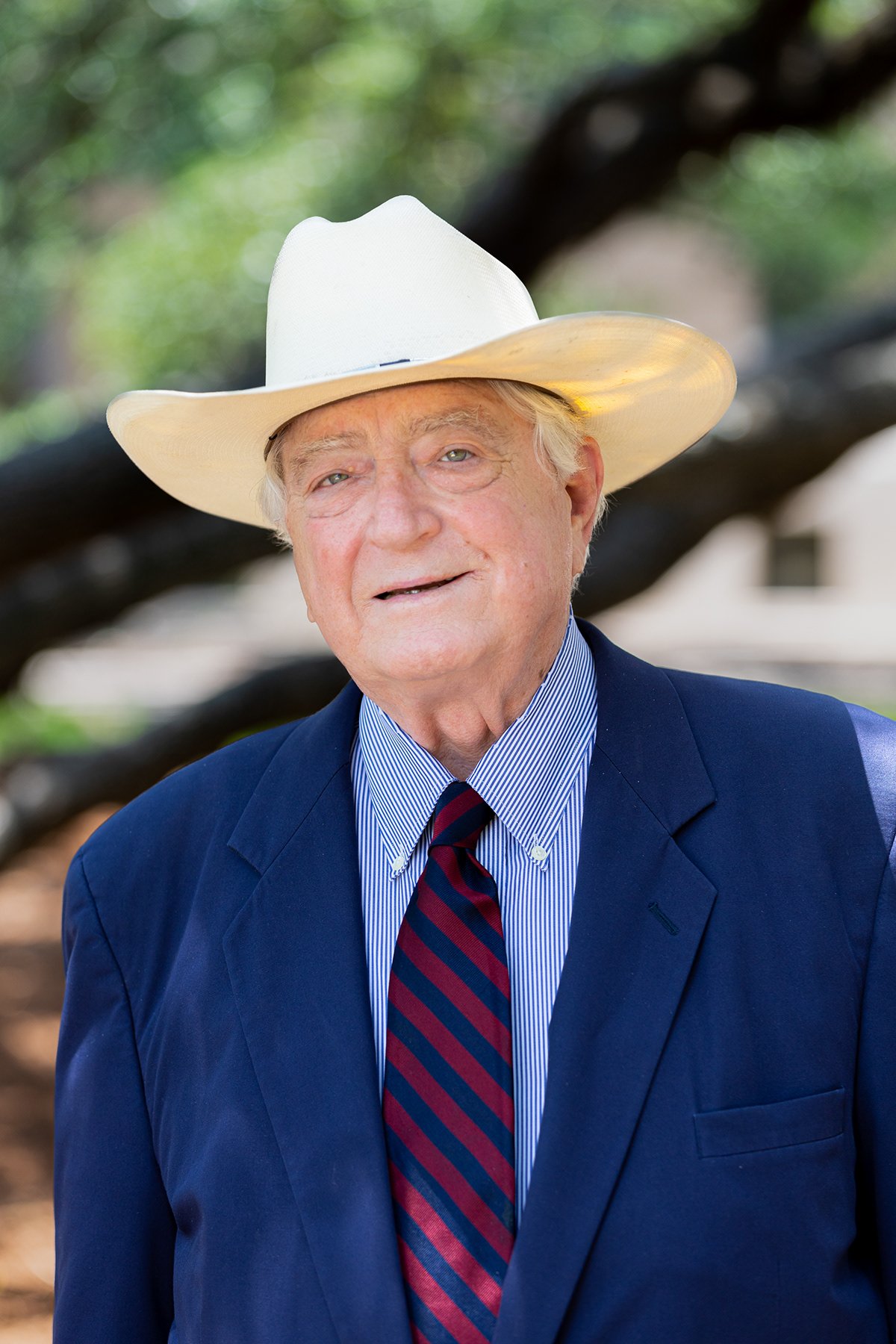 Older man with light skin, gray hair, and blue eyes wearing a white cowboy hat, a blue suit, a striped blue and white shirt, and a red and blue striped tie, standing outdoors with a blurred background of trees.