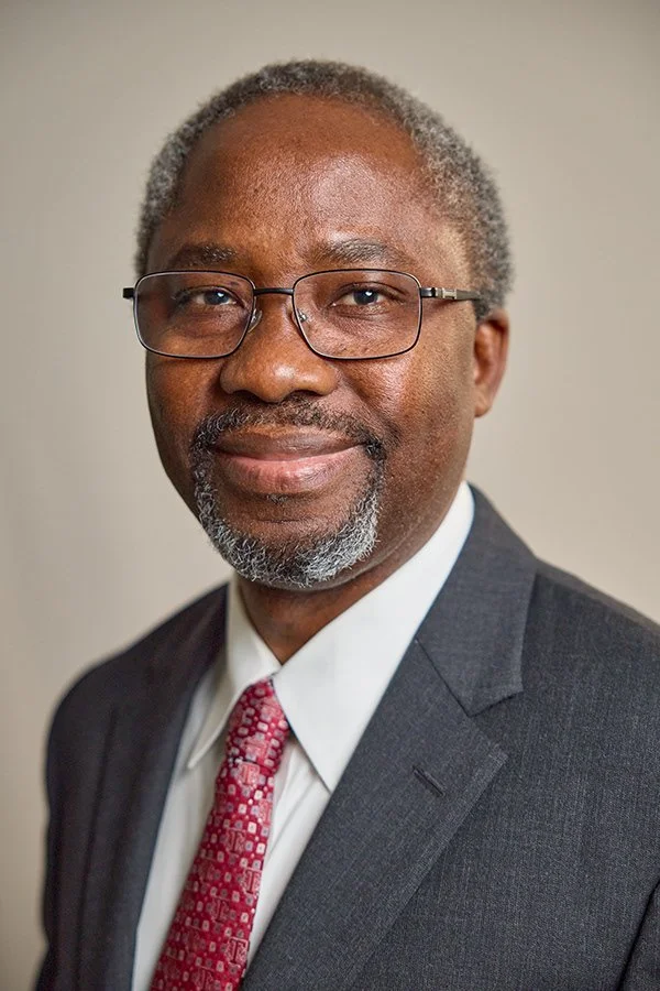 Portrait of a middle-aged African American man with gray hair and glasses, wearing a dark suit, white shirt, and red patterned tie.
