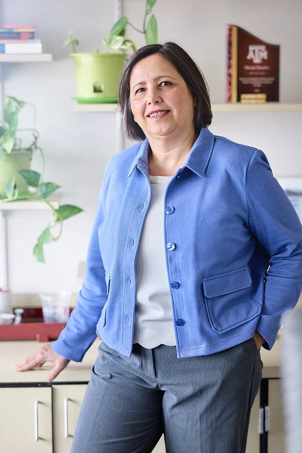 A woman stands in an office or classroom environment, smiling at the camera. She wears a blue jacket over a white top and gray striped pants, with one hand resting on a desk or counter behind her.