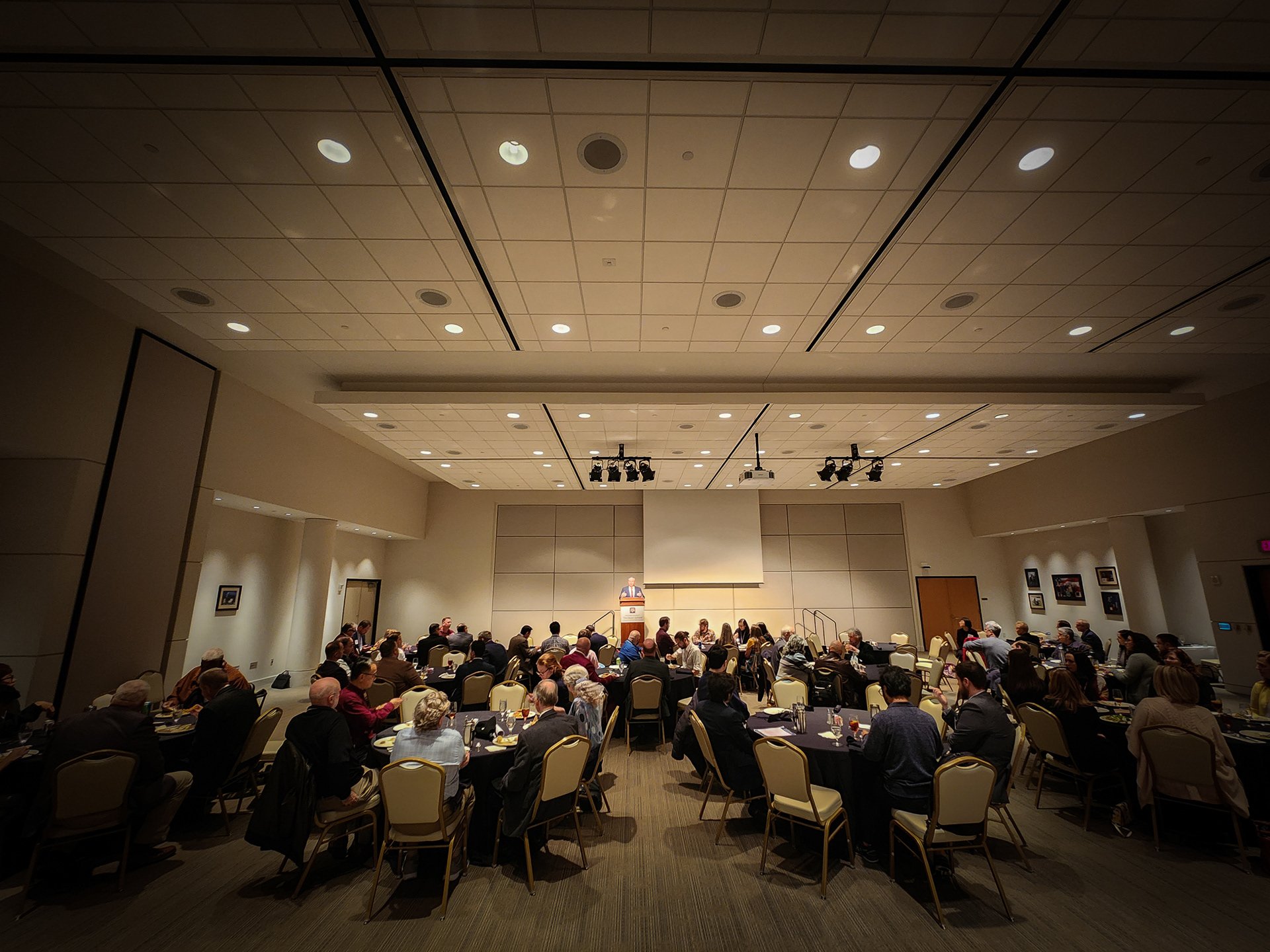 A large conference room filled with round tables and people attending a formal meeting or event. A presenter stands at a podium at the front of the room under ceiling lights, with a projection screen behind them. Attendees are listening and taking no