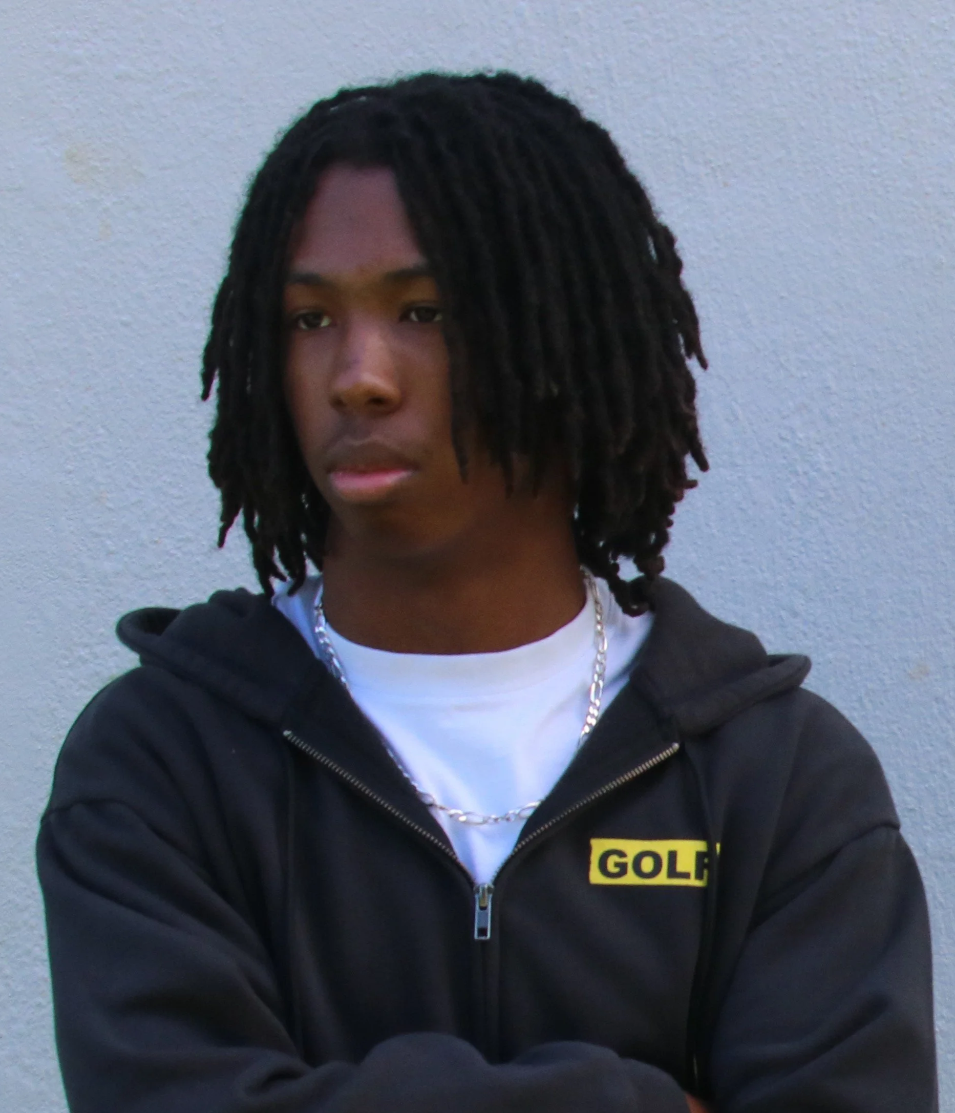 A young person with curly dreadlocks standing against a plain wall, wearing a black hoodie with a yellow patch reading 'GOLF' and a white shirt underneath, accessorized with a chain necklace.