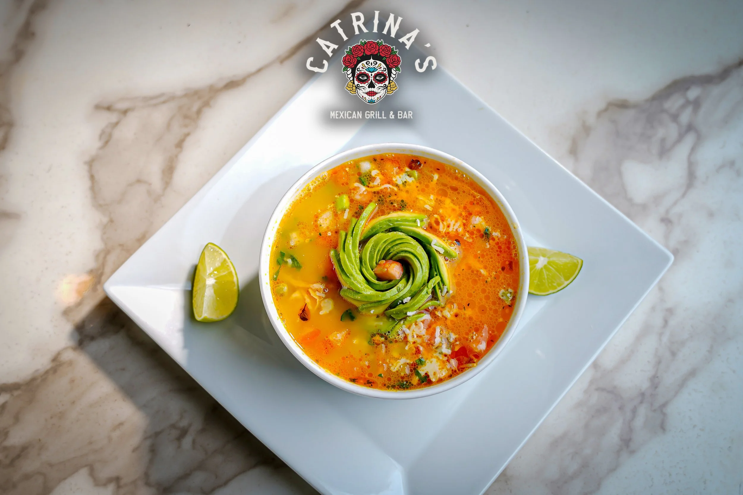A bowl of vibrant yellow-orange soup garnished with sliced avocado and lime wedges on a white plate, with a marble countertop in the background and Fortatina Mexican Grill & Bar logo at the top.