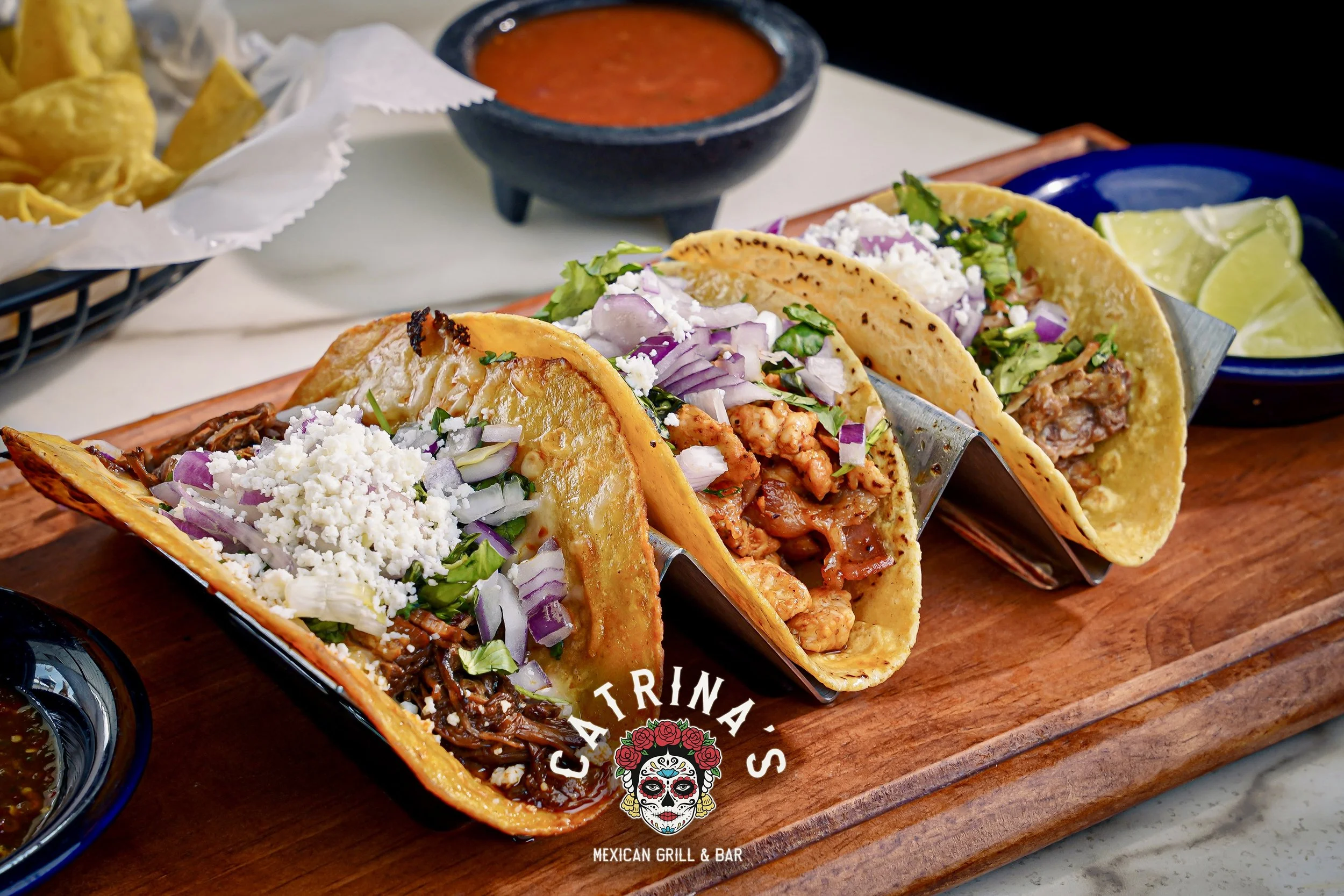 Three tacos filled with meat, lettuce, shredded cheese, chopped onions, and other toppings on a wooden serving tray with lime wedges and salsa in bowls.