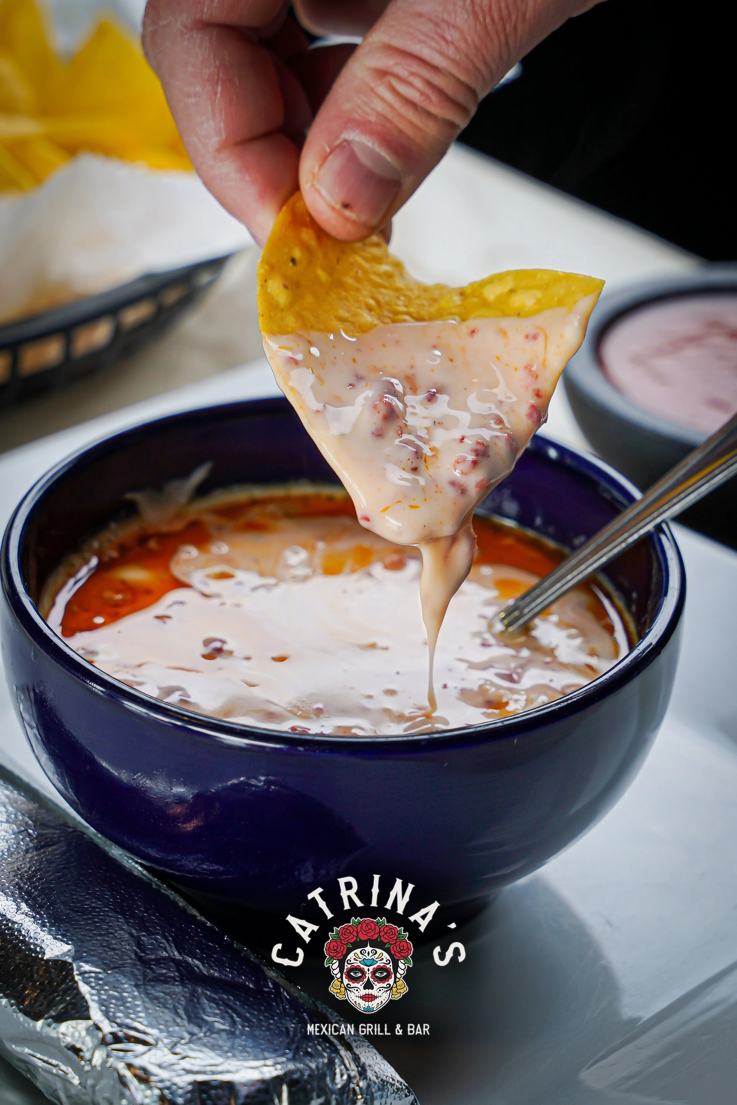 Close-up of a hand dipping a tortilla chip into a bowl of creamy queso cheese at a Mexican restaurant, with other bowls of sauces in the background.
