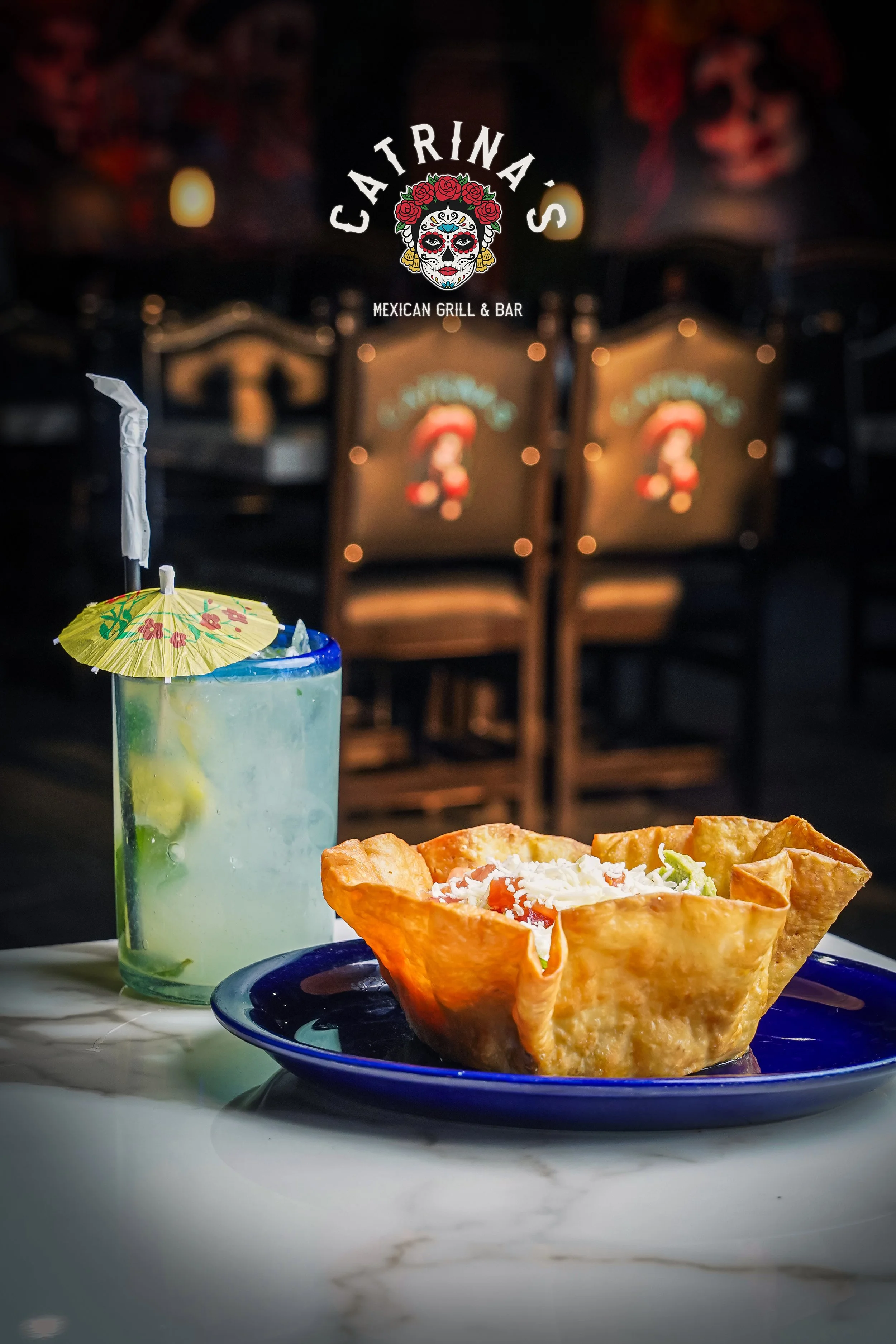 A plate of Mexican food with a crispy taco filled with lettuce, tomato, and cheese, and a margarita cocktail with a small decorative umbrella, set on a marble table with a Mexican Grill & Bar logo in the background.