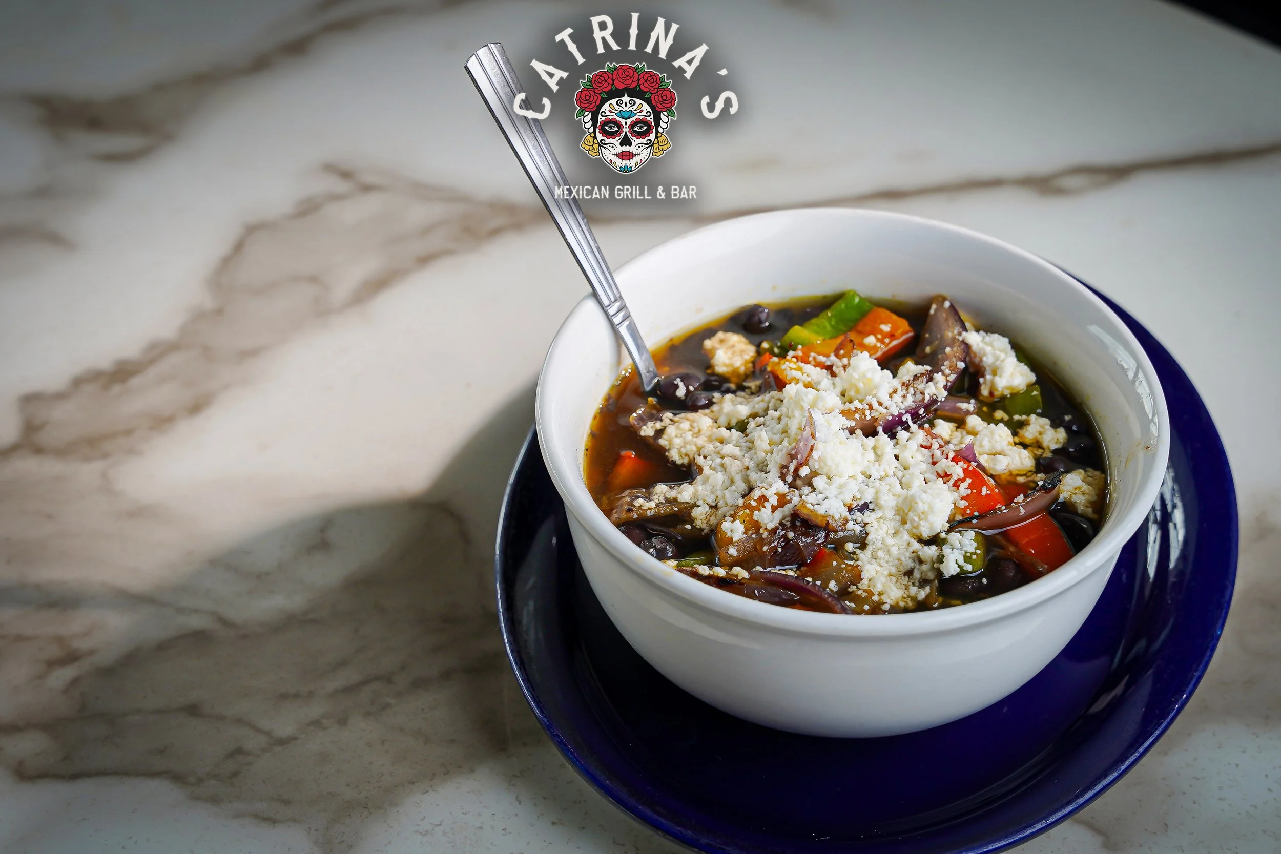 A bowl of black bean soup with vegetables and cheese, placed on a blue plate on a white marble countertop.