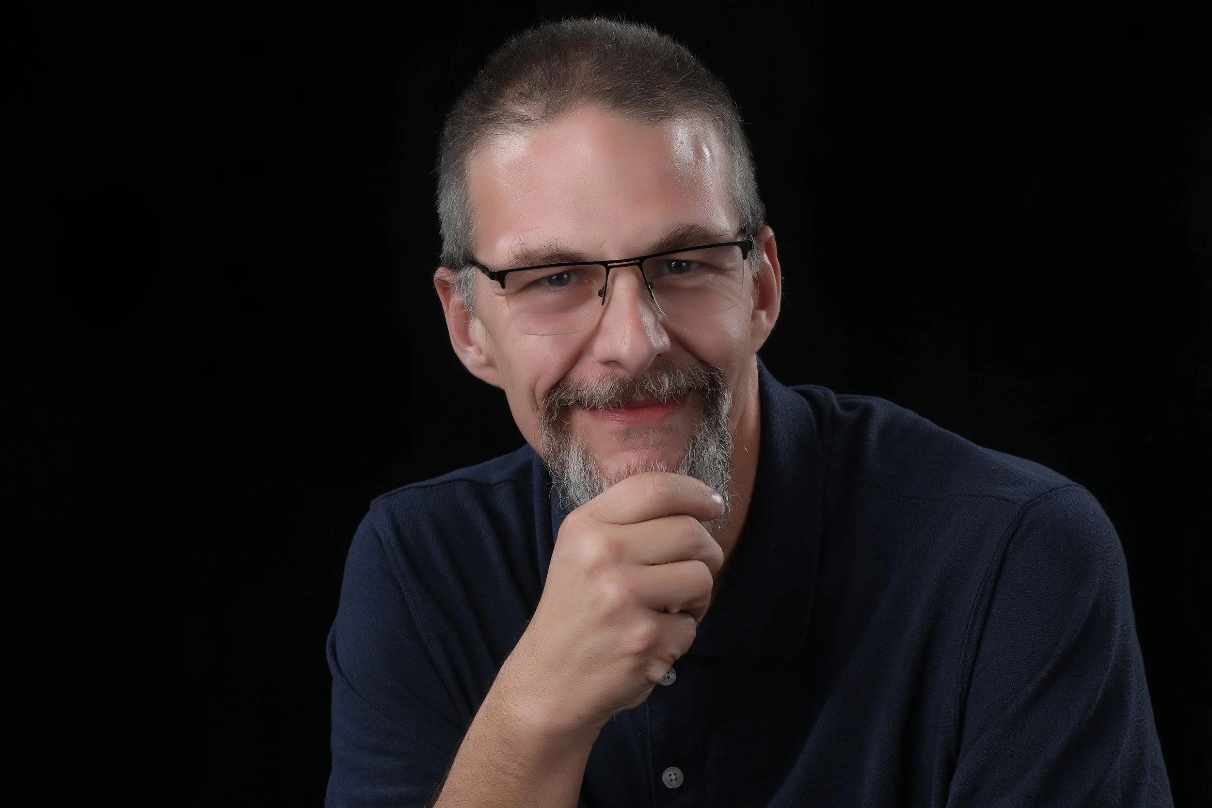 A man with glasses, a beard, and short hair, smiling and holding his chin with his hand, wearing a dark blue shirt, against a black background.