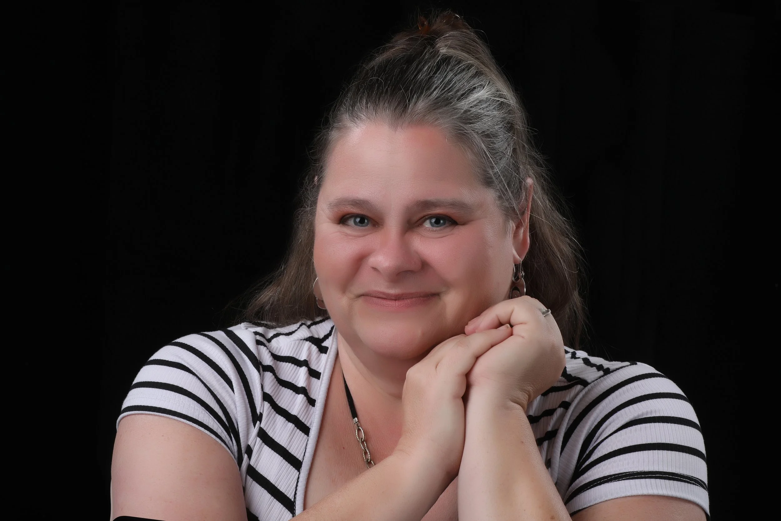 A woman with blonde hair and blue eyes, wearing a striped shirt and silver jewelry, smiles softly with her hands resting on her chin against a black background.