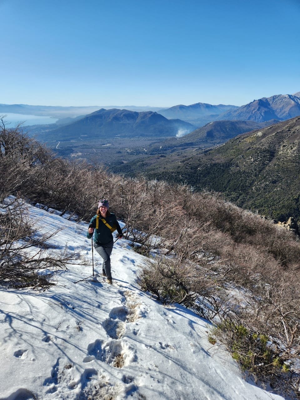 Carolyn Noelke, CCC-SLP, hiking on a snowy trail with mountains and a blue sky in the background.