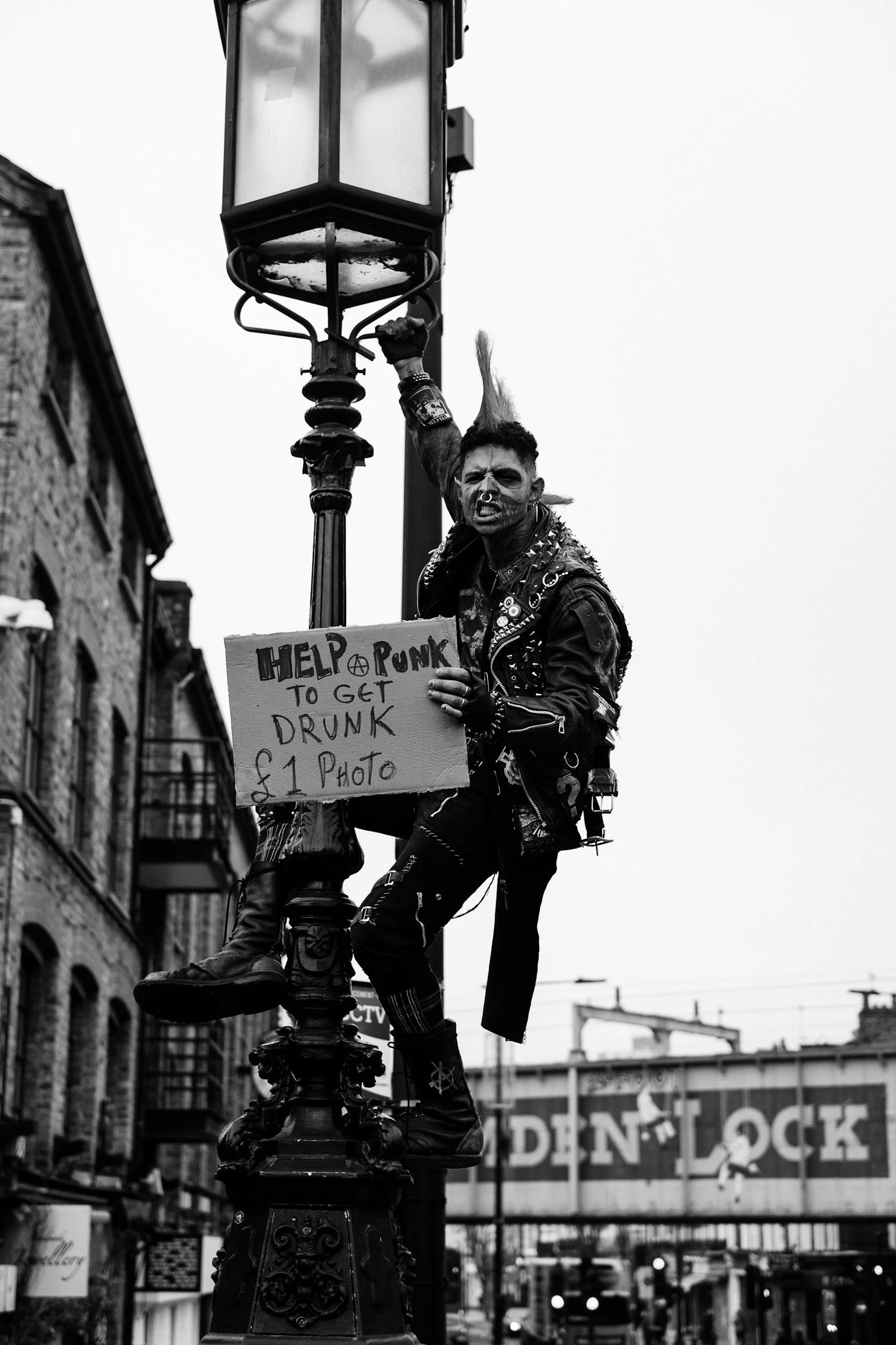 A person with a punk appearance, including spiked hair, face paint, and studs on their clothing, is holding onto a streetlamp while standing on it. They are also holding a cardboard sign that reads 'Help Punk to get Drunk & 1 Photo' in an urban area, with buildings and a bridge visible in the background.