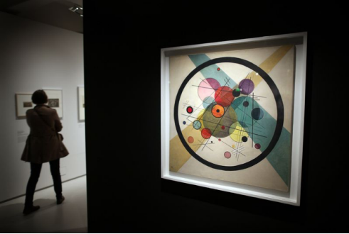 A woman viewing an abstract geometric painting in an art gallery, featuring circles and intersecting shapes in vibrant colors.