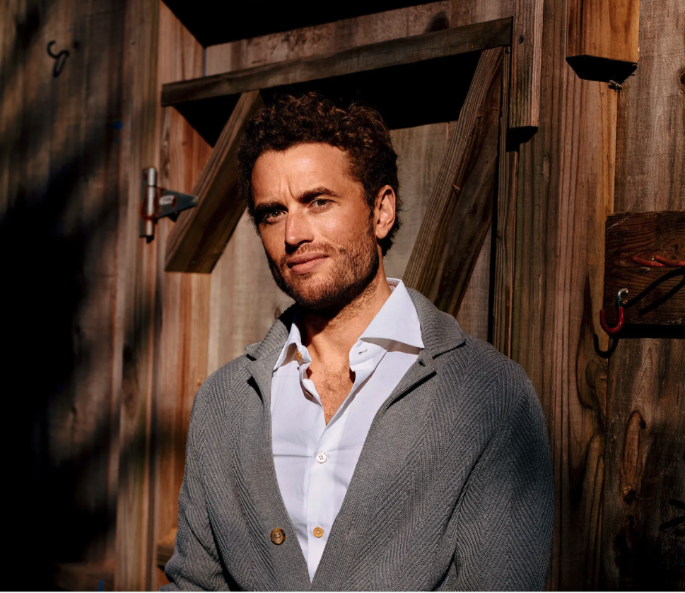 A man with curly hair and a beard, wearing a white shirt and a gray cardigan, standing in front of a wooden wall with some tools and structures visible.